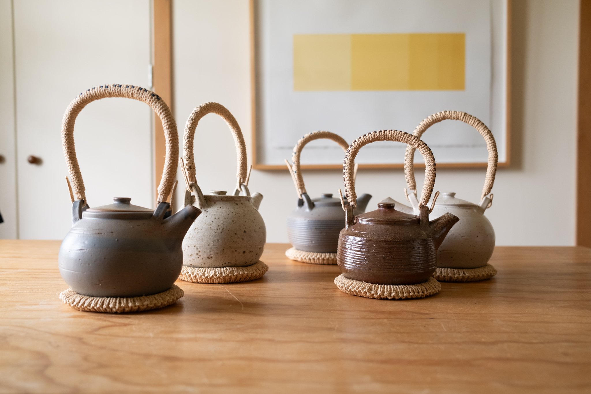 Six small teapots on a wooden table, each resting on woven coasters with braided handles.