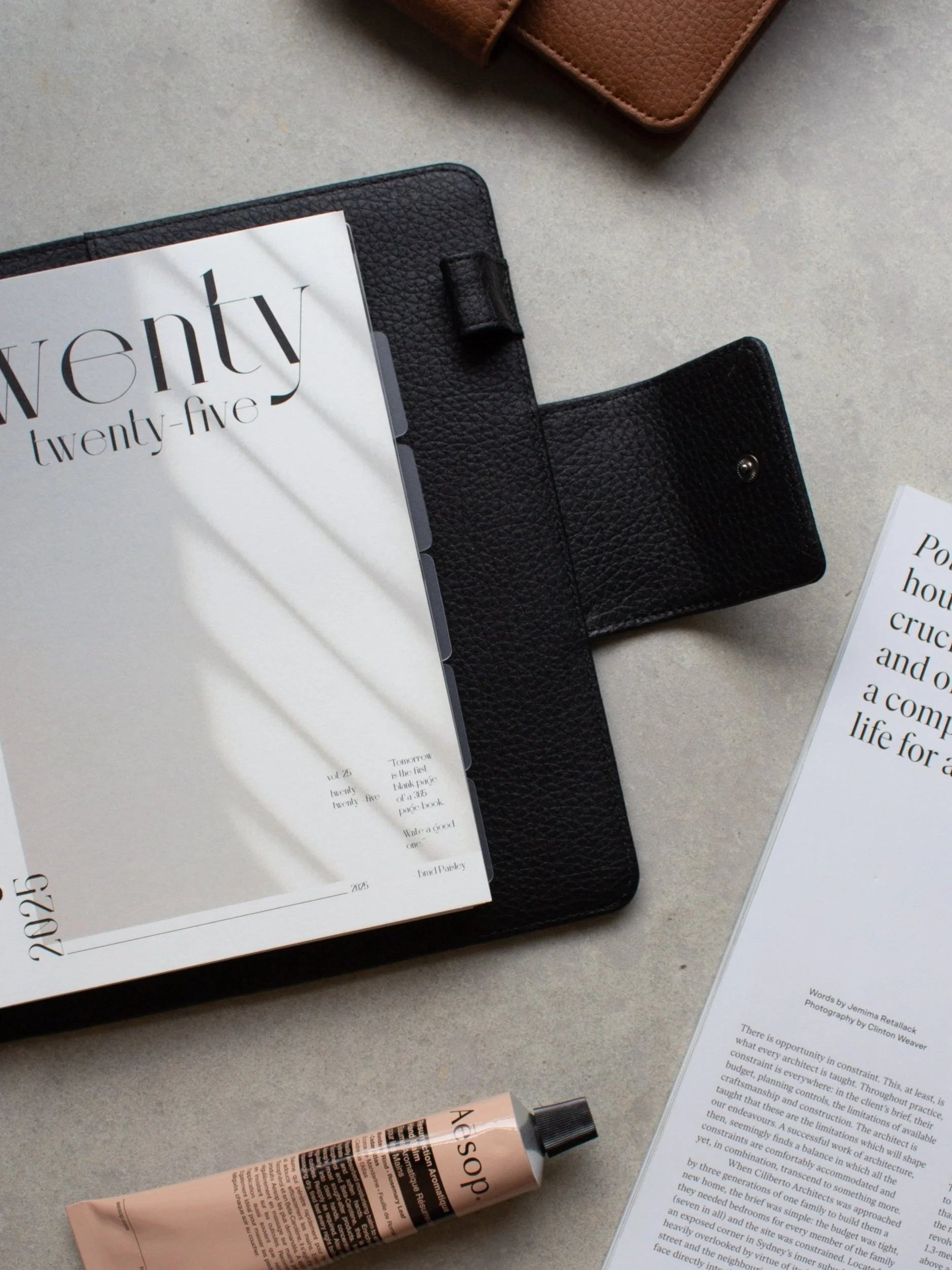 Flat lay of a black leather planner, a calendar or magazine with the year 2025 on it, a printed article, and a tube of skincare or hand cream on a light gray surface.