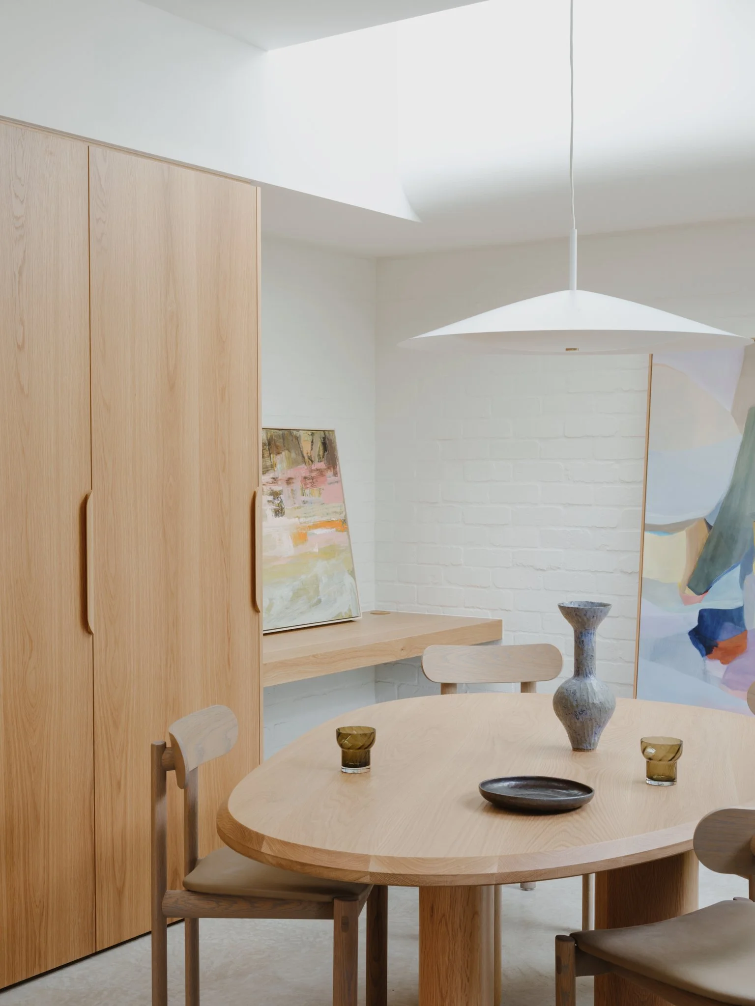 A dining room with a round wooden table, four wooden chairs, a white hanging lamp, a large wooden wardrobe, a small painting, two vases, and a tray, with a white brick wall and a colorful abstract artwork.