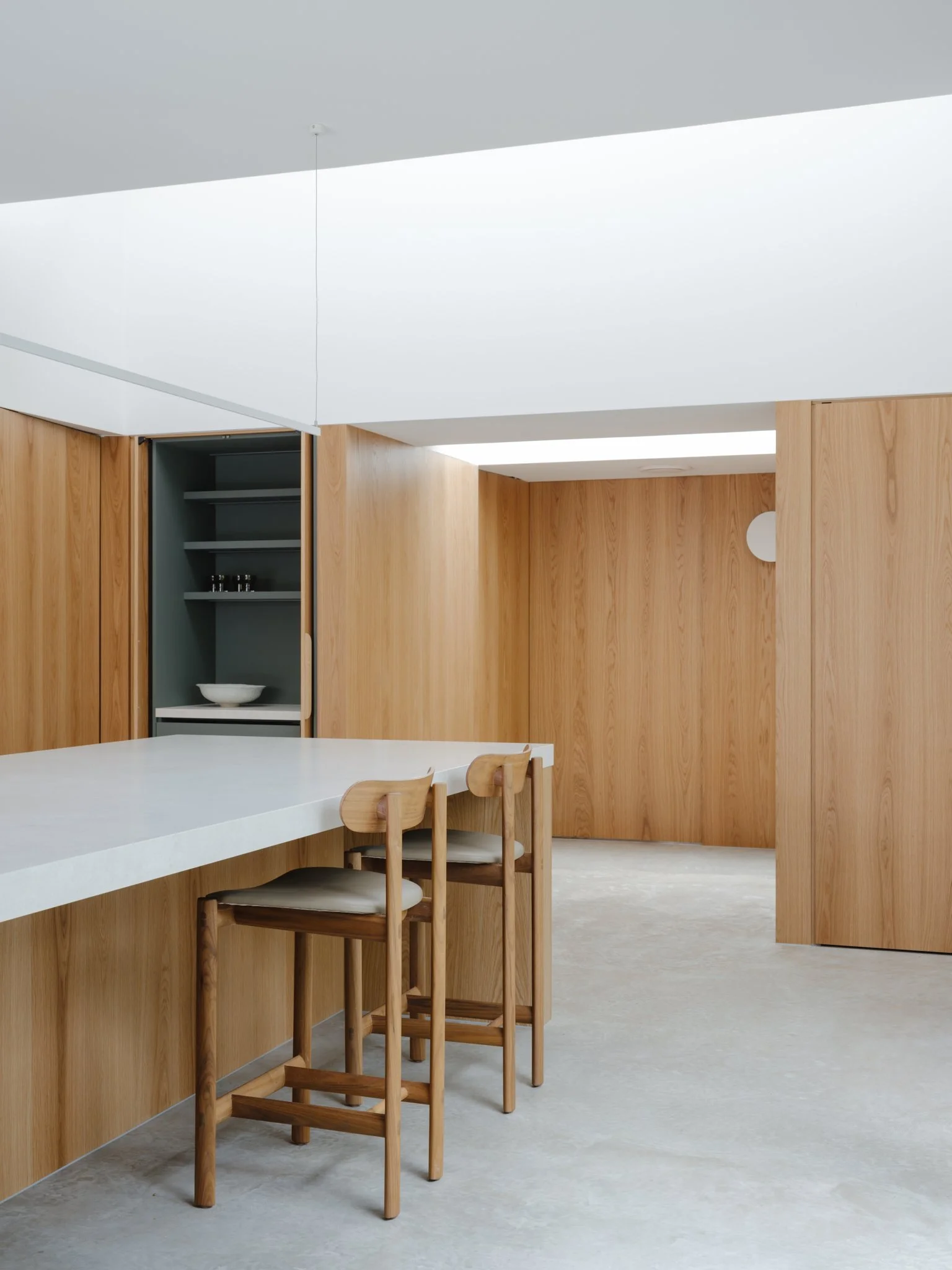 A minimalist kitchen with light wood paneling, a white countertop, and three wooden bar stools with cushions.