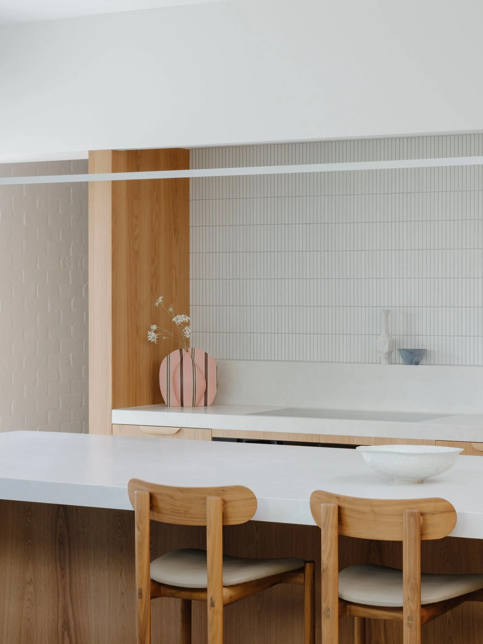 Modern kitchen with white countertops, wooden chairs, a decorative pink and white vase with flowers, and minimalistic decor.