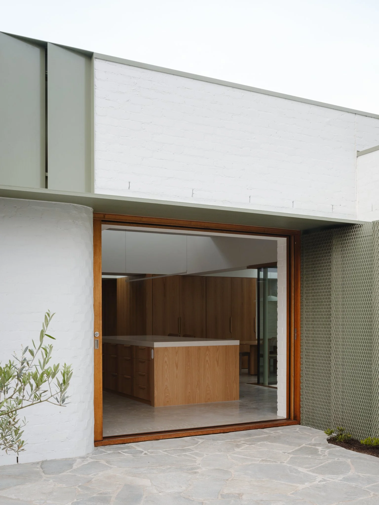 A modern house with a large sliding glass door that opens to an outdoor stone patio. The interior visible through the door features wooden kitchen cabinets and an island.