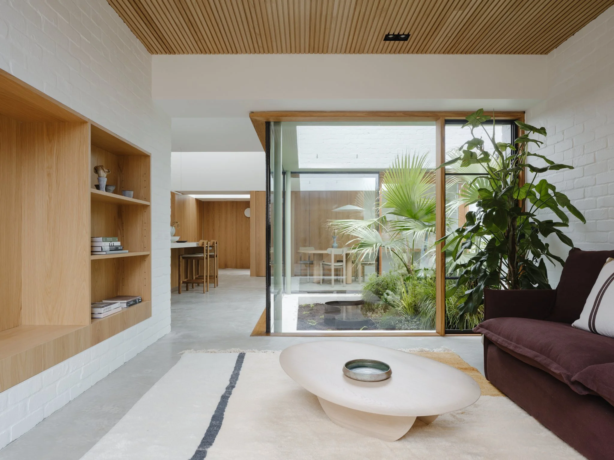 Modern living room with a large glass window overlooking indoor plants, a maroon sofa with a pillow, a white rug with black lines, and a round white coffee table with a small tray.
