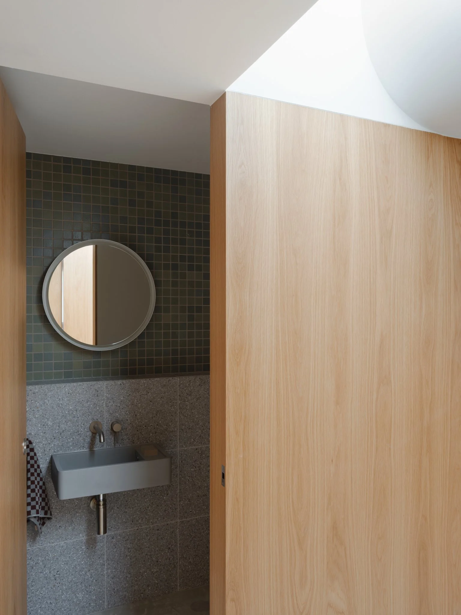 Small bathroom with gray sink, round mirror, dark green tiled wall, light wood partitions, and natural light from skylight.