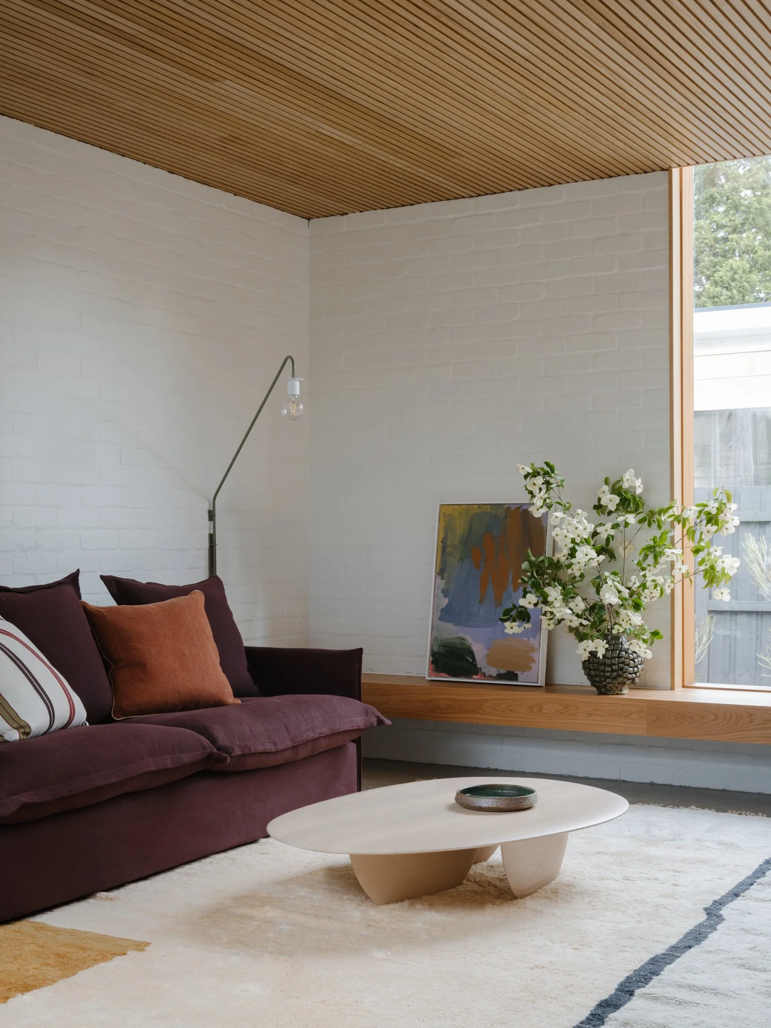 Modern living room with a purple sofa, a wooden coffee table, a large window, a white brick wall, a floor lamp with a bare bulb, an abstract painting, and a floral display in a black vase.