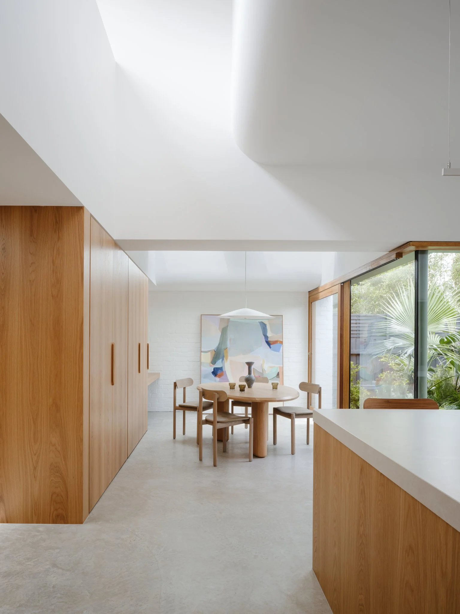 Modern dining area with a round wooden table, four matching chairs, a large abstract painting on a white brick wall, and large windows with greenery outside.