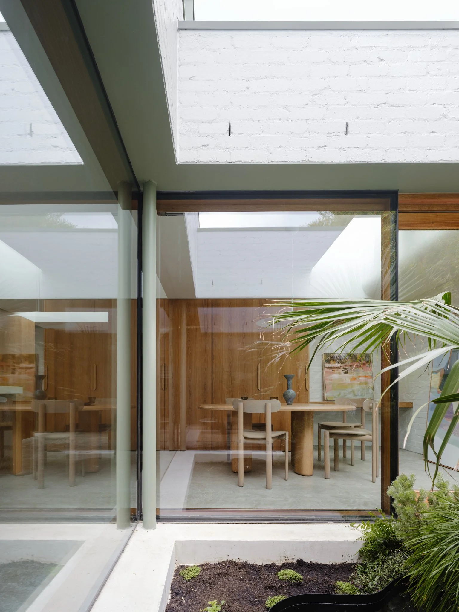 Interior view of a modern house with wooden walls, large glass windows, a dining table with chairs, and plants outside.