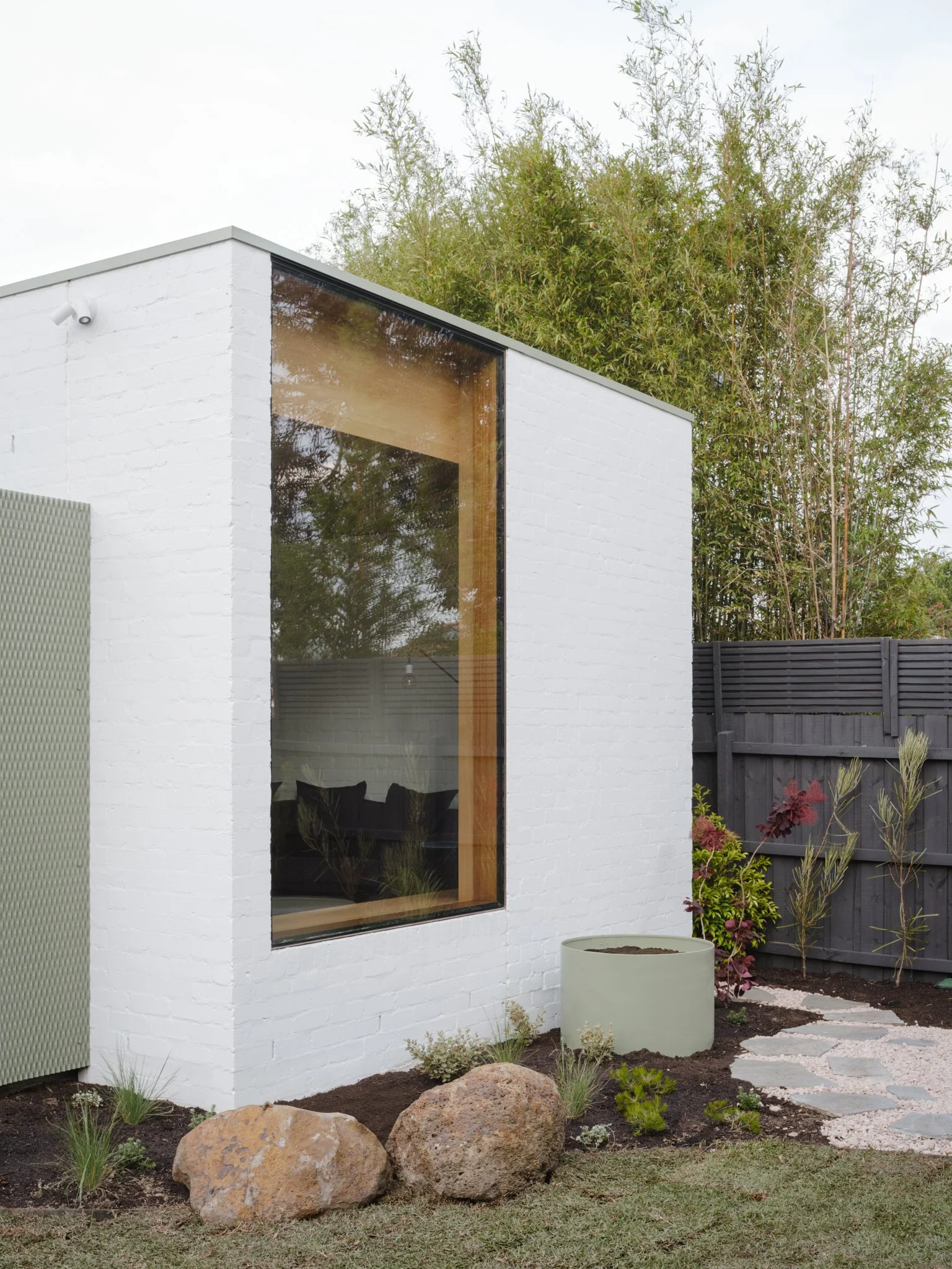 Modern white building with large corner window, black fence, plant bed with rocks and plants, stepping stones pathway, outdoor greenery