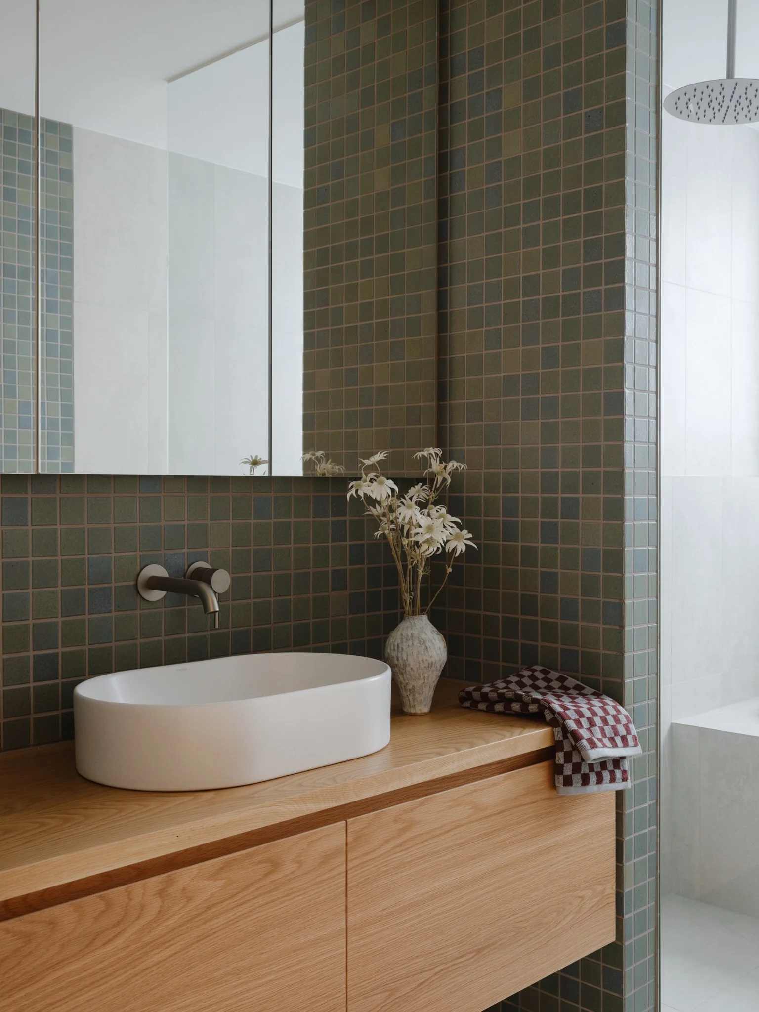 Modern bathroom with a wooden vanity, oval white sink, wall-mounted faucet, a vase with white flowers, and a checkered towel. Green mosaic tiles on the wall and a glass shower with a rain showerhead.