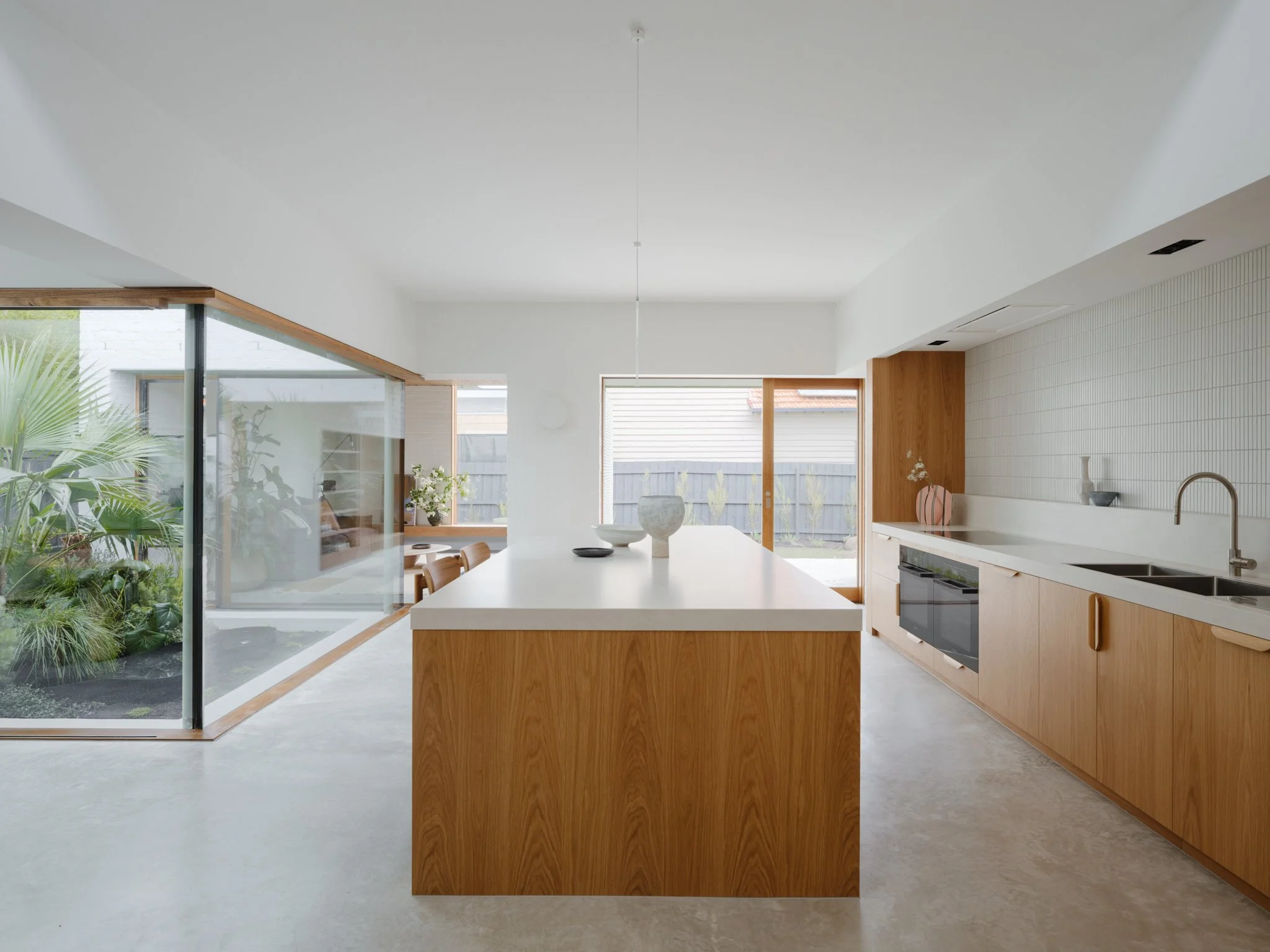 Modern kitchen with wooden cabinets, white countertop, and large glass windows overlooking a garden.
