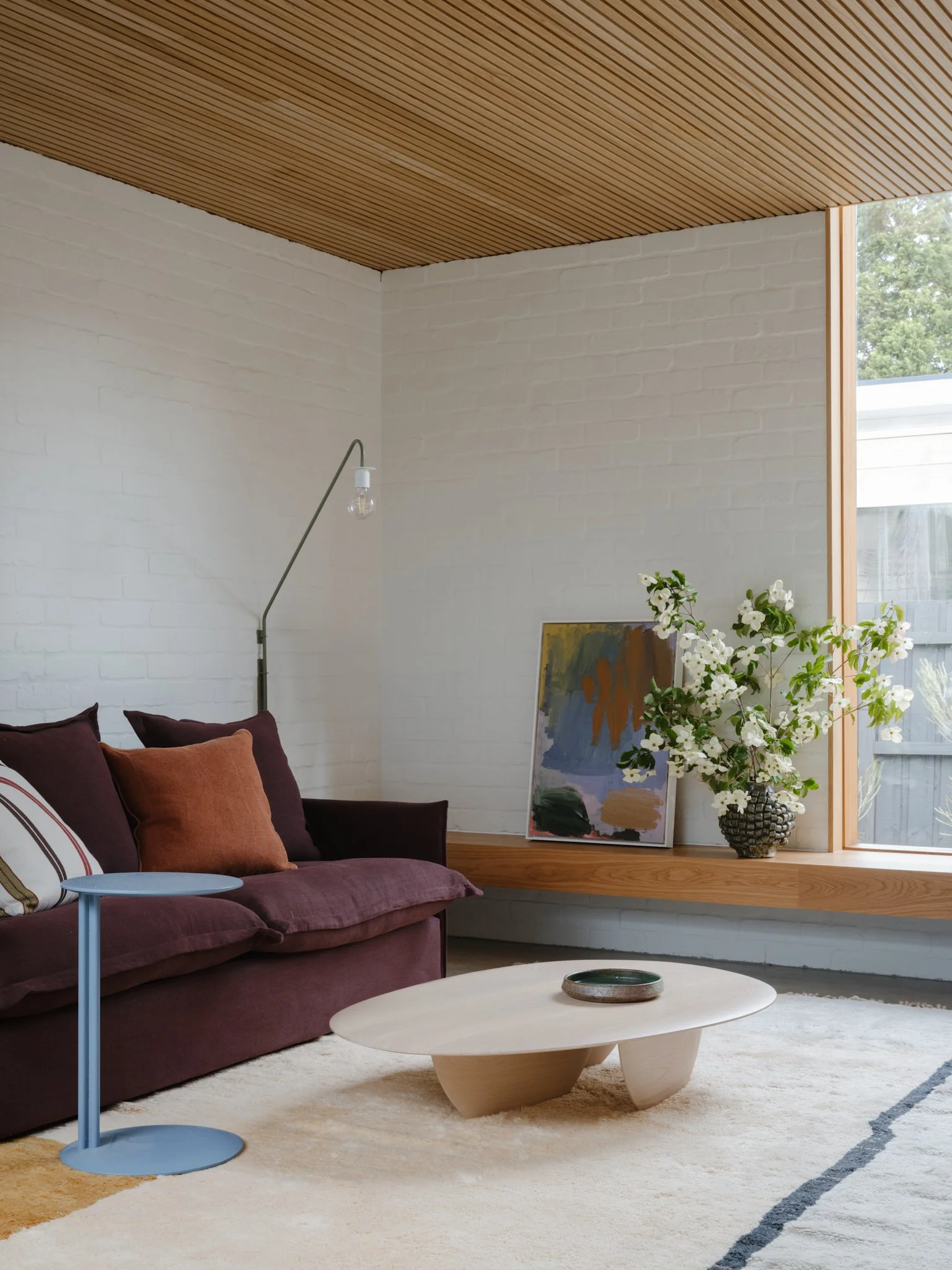 Living room with a maroon sofa, a light-colored modern coffee table, a side table, a floor lamp, a white brick wall, a large window with a ledge, a vase with white flowers, and an abstract painting.