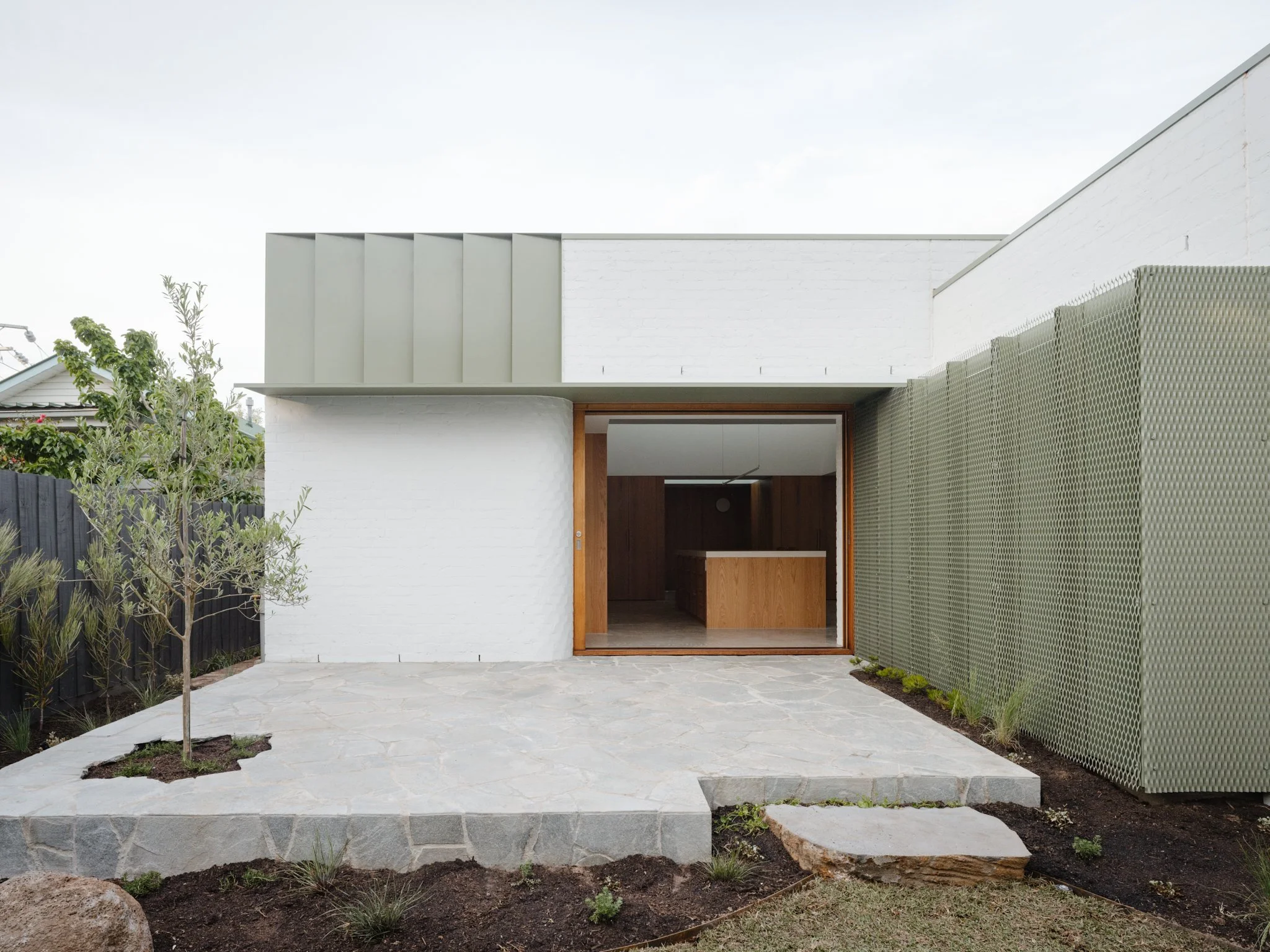 Modern house with a white brick exterior, wooden sliding door, stone patio, and green privacy screen on the side.