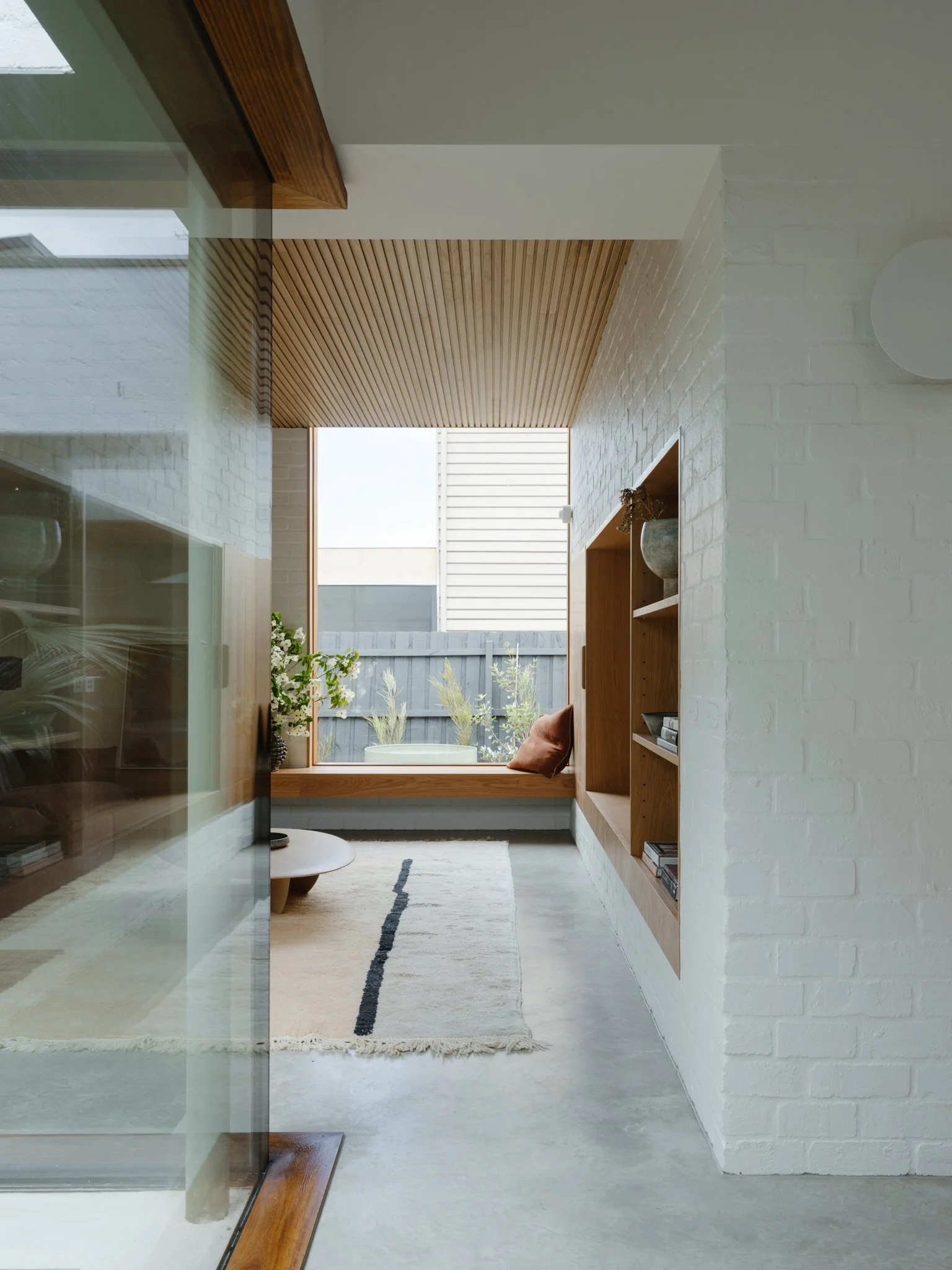 A modern interior room with white brick walls, wooden ceiling, large window seat with pillows, and a view of a fenced backyard with plants.