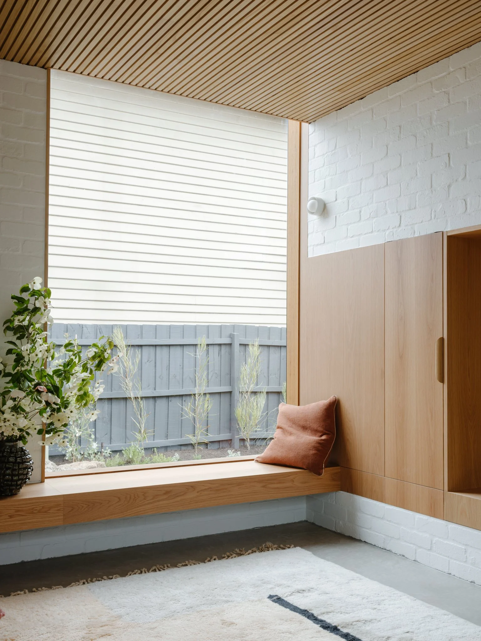 Interior corner with a wooden window seat, a brown pillow, a potted plant, and a view of a backyard with a wooden fence and small plants.
