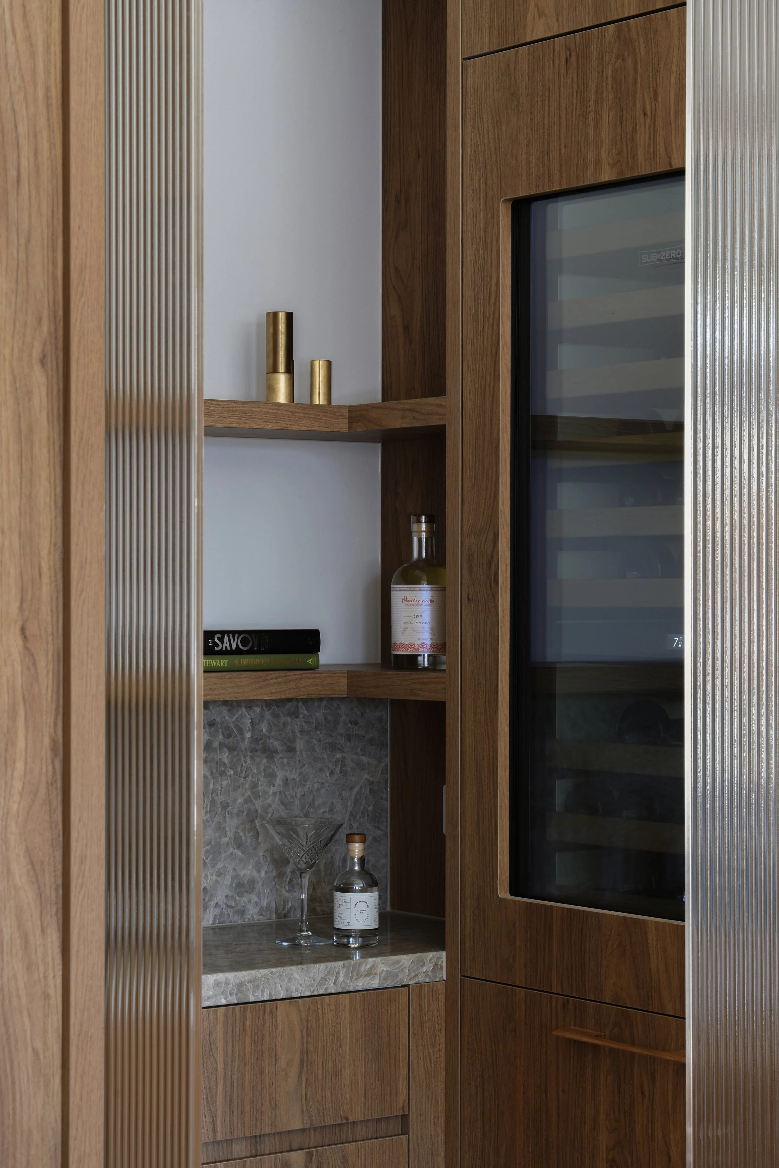 View of a modern kitchen cabinet with wooden and metal finishes, containing a glass-front wine cooler, shelves with decorative gold vases, a couple of books, a bottle of alcohol, and a marble countertop with a martini glass and a small bottle.