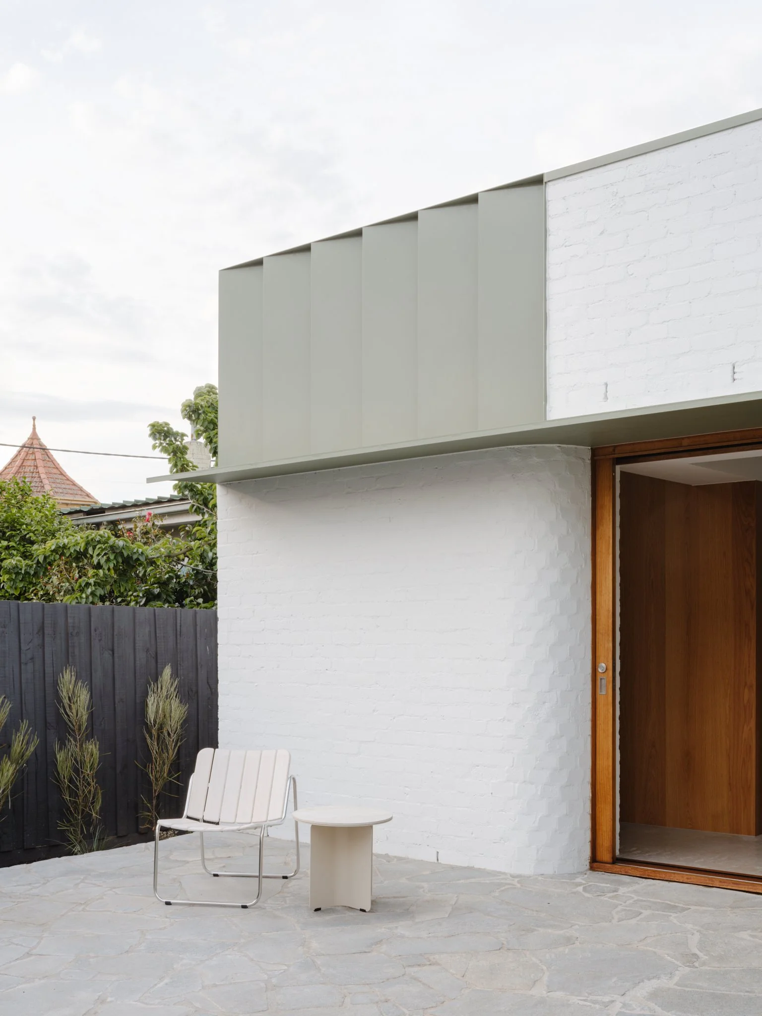 Modern house with a white brick wall, sliding wooden door, outdoor patio with a white chair and small table, black fence, and greenery in the background.