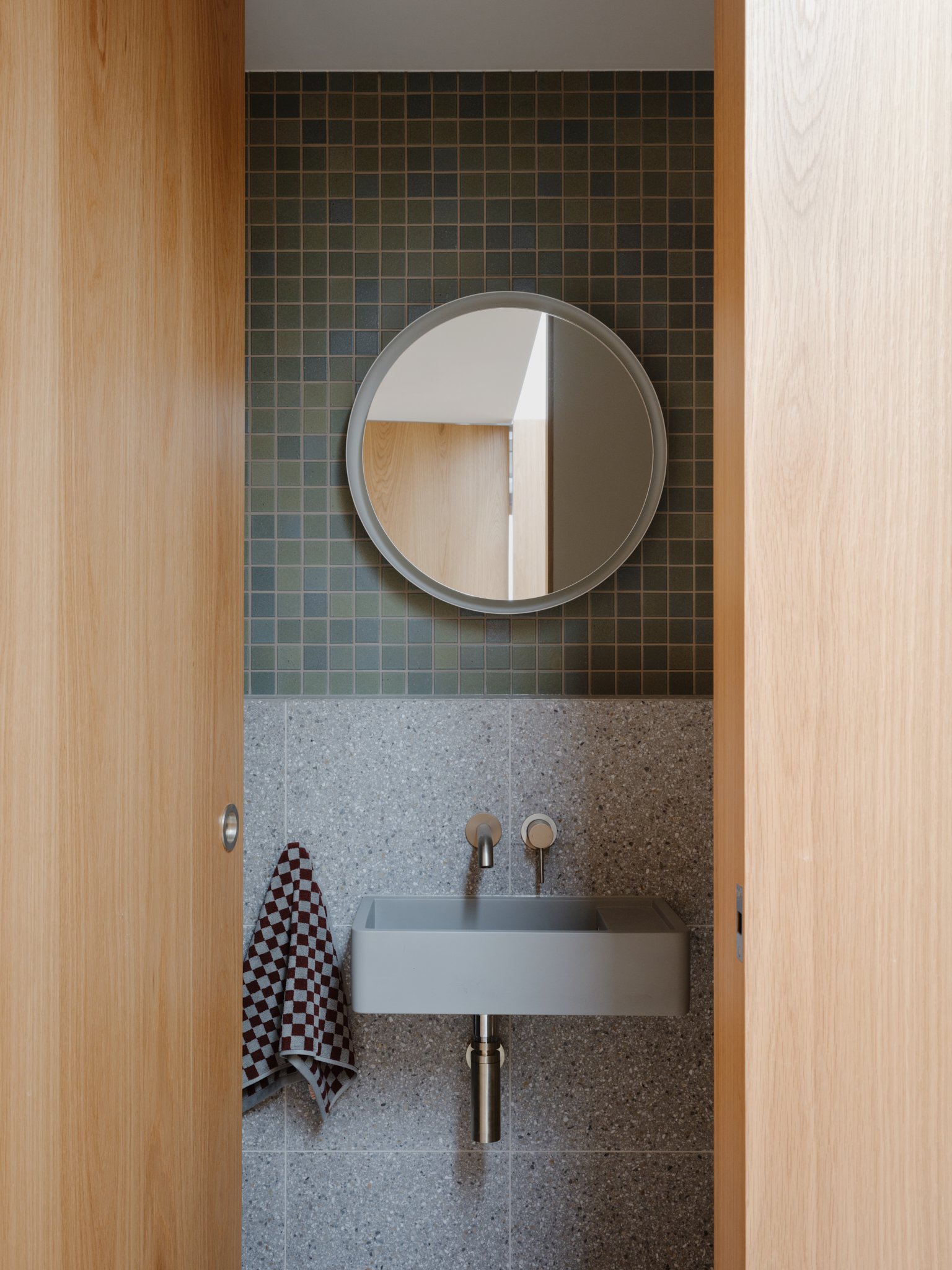 Small bathroom with a gray round mirror above a rectangular wall-mounted sink, checkered towel hanging on a hook, and dark square tiles on the wall behind the mirror, with light wooden doors on each side.