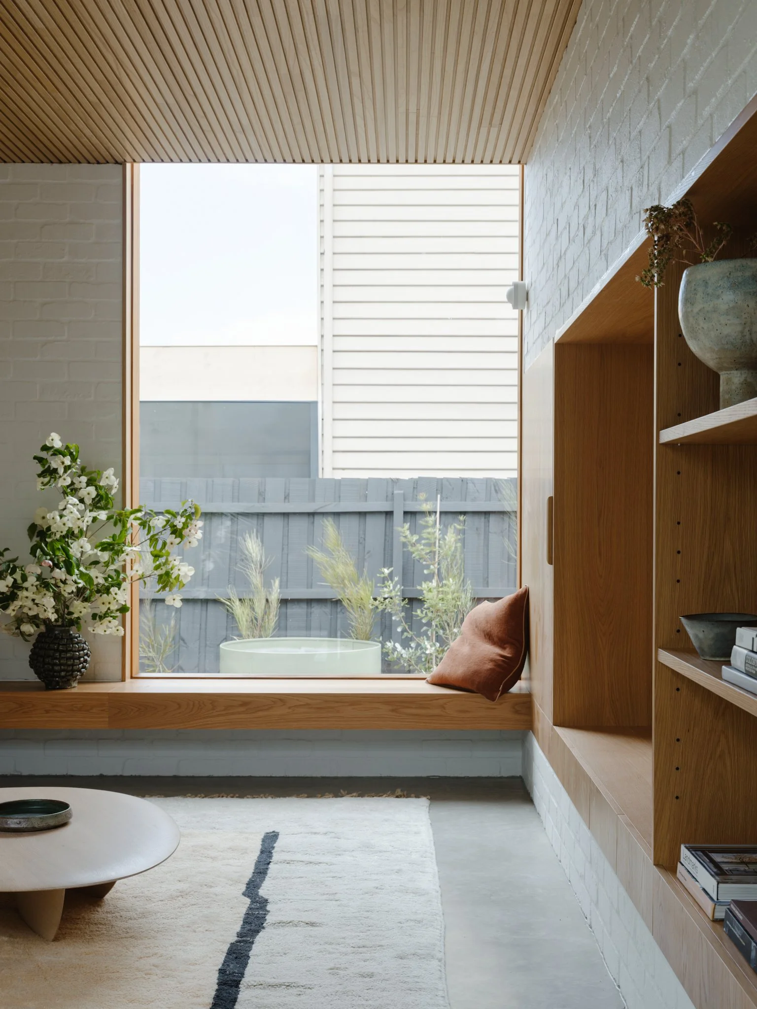 Modern interior with large window seat, wooden shelves, potted plants, and a white rug.