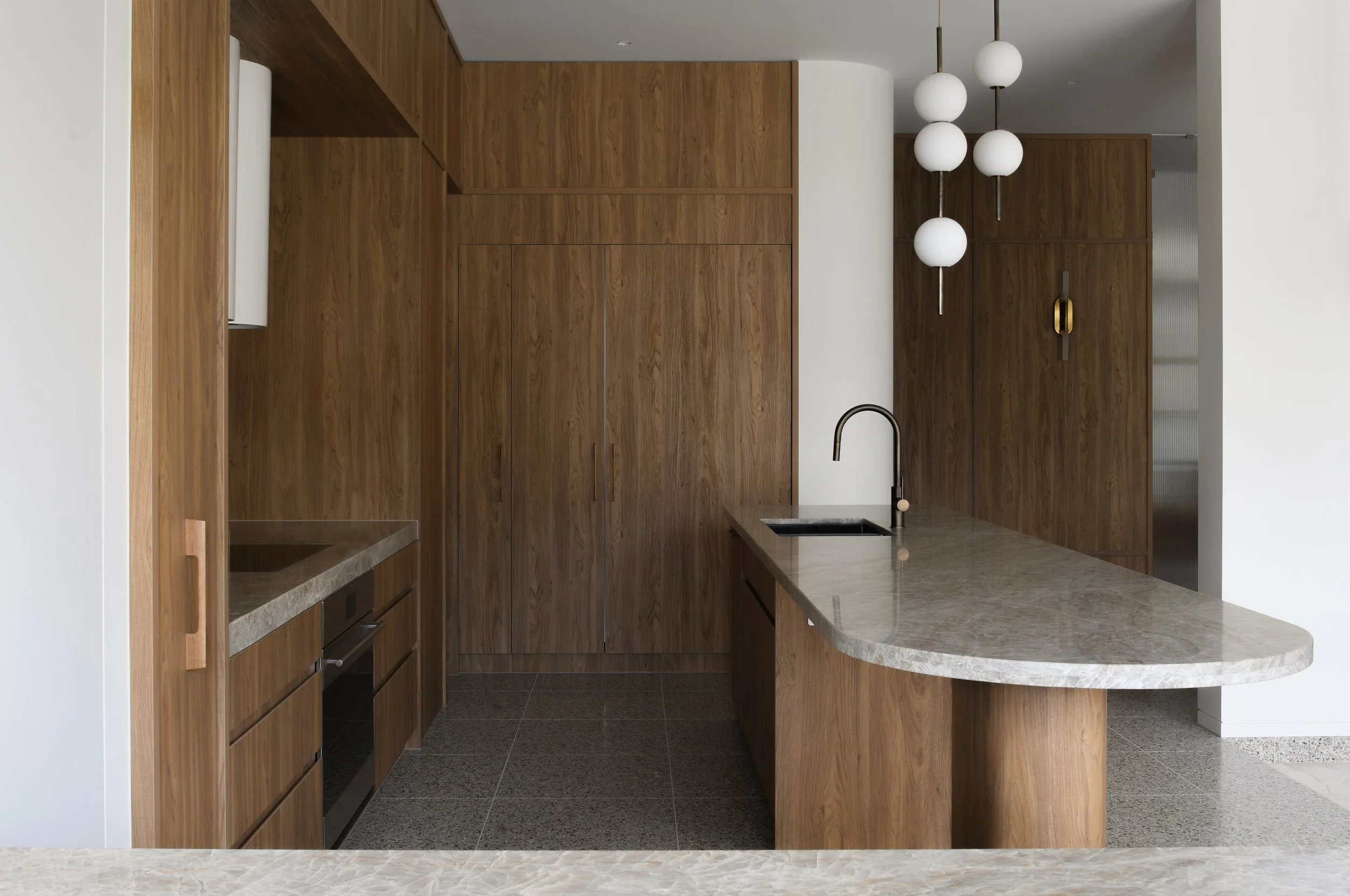 Modern kitchen with wooden cabinets, a marble countertop island, pendant white globe lights, and a black faucet.