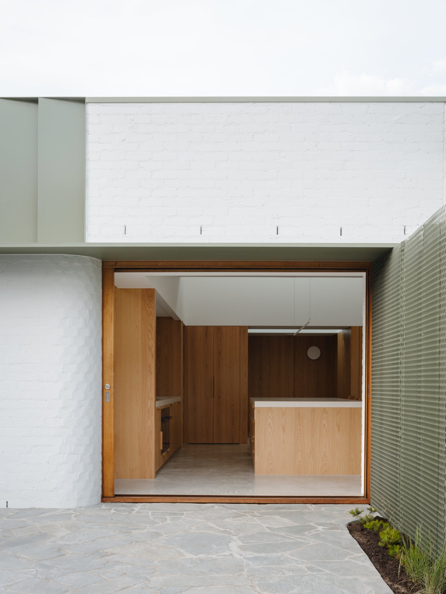 View of a modern interior kitchen with wooden cabinets, open doorway, and white brick wall outside