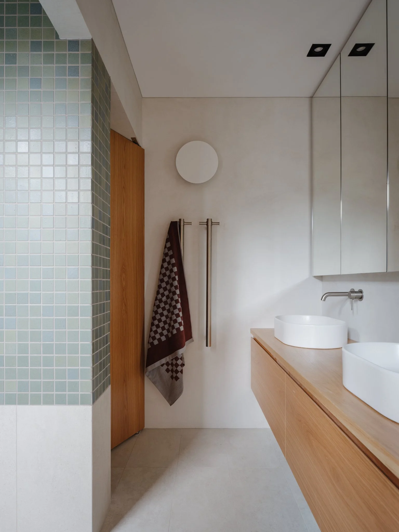 Modern bathroom with wooden vanity, two white vessel sinks, black faucet, mirrored cabinets, circular wall light, and a towel hanging on a rack next to a wooden door.