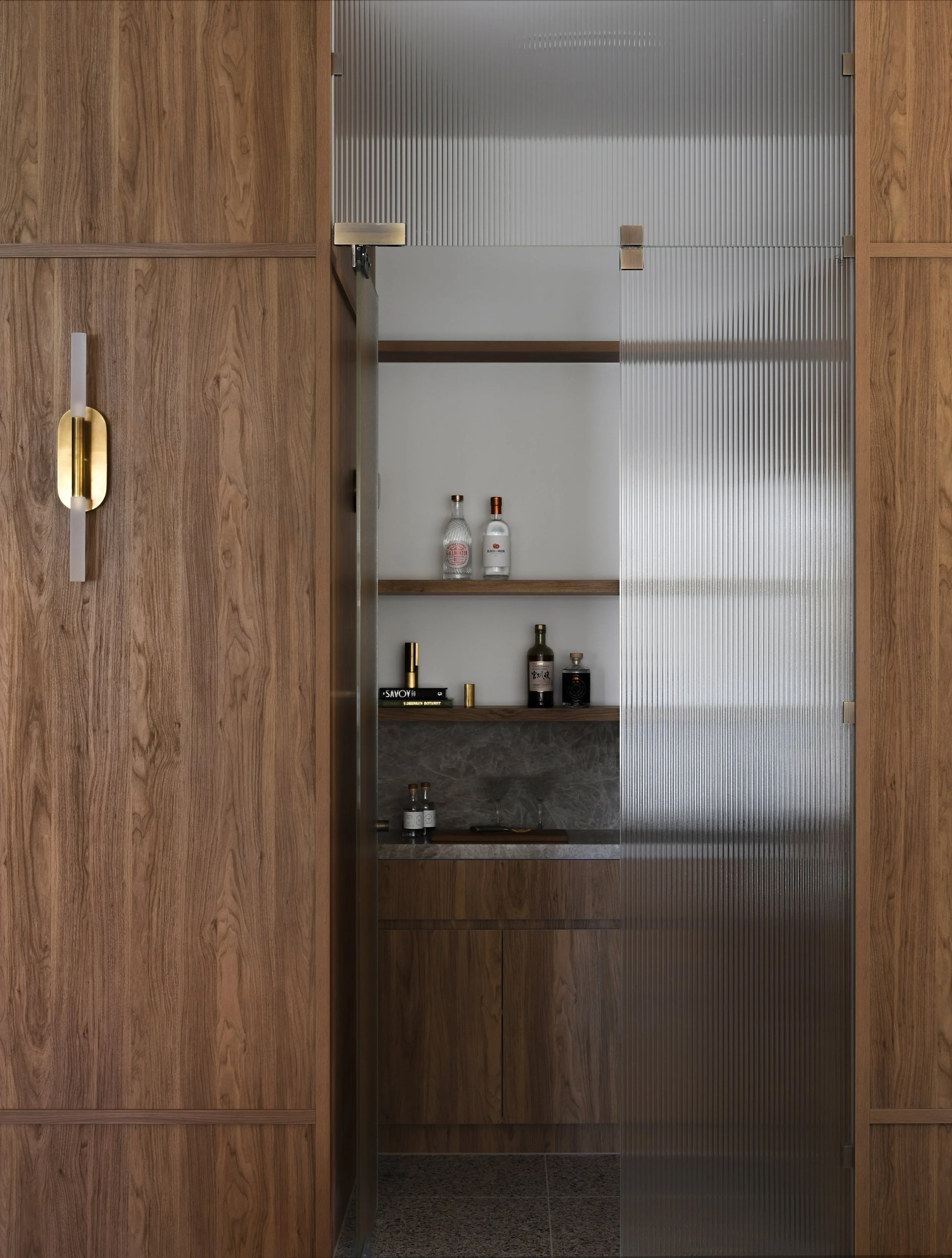 View of a built-in wooden cabinet with a frosted glass sliding door, revealing shelves with bottles and small decorative items inside.
