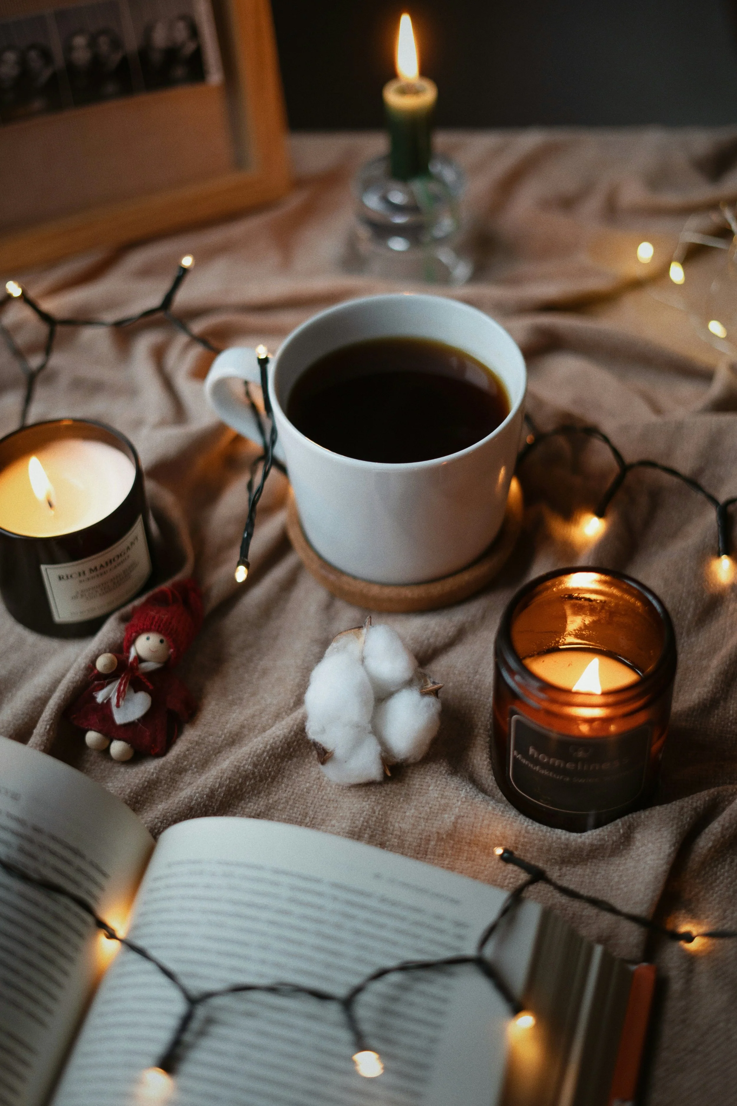 A cozy scene featuring an open book, a cup of coffee, lit candles, fairy lights, and a small decorative doll, creating a warm and inviting atmosphere.