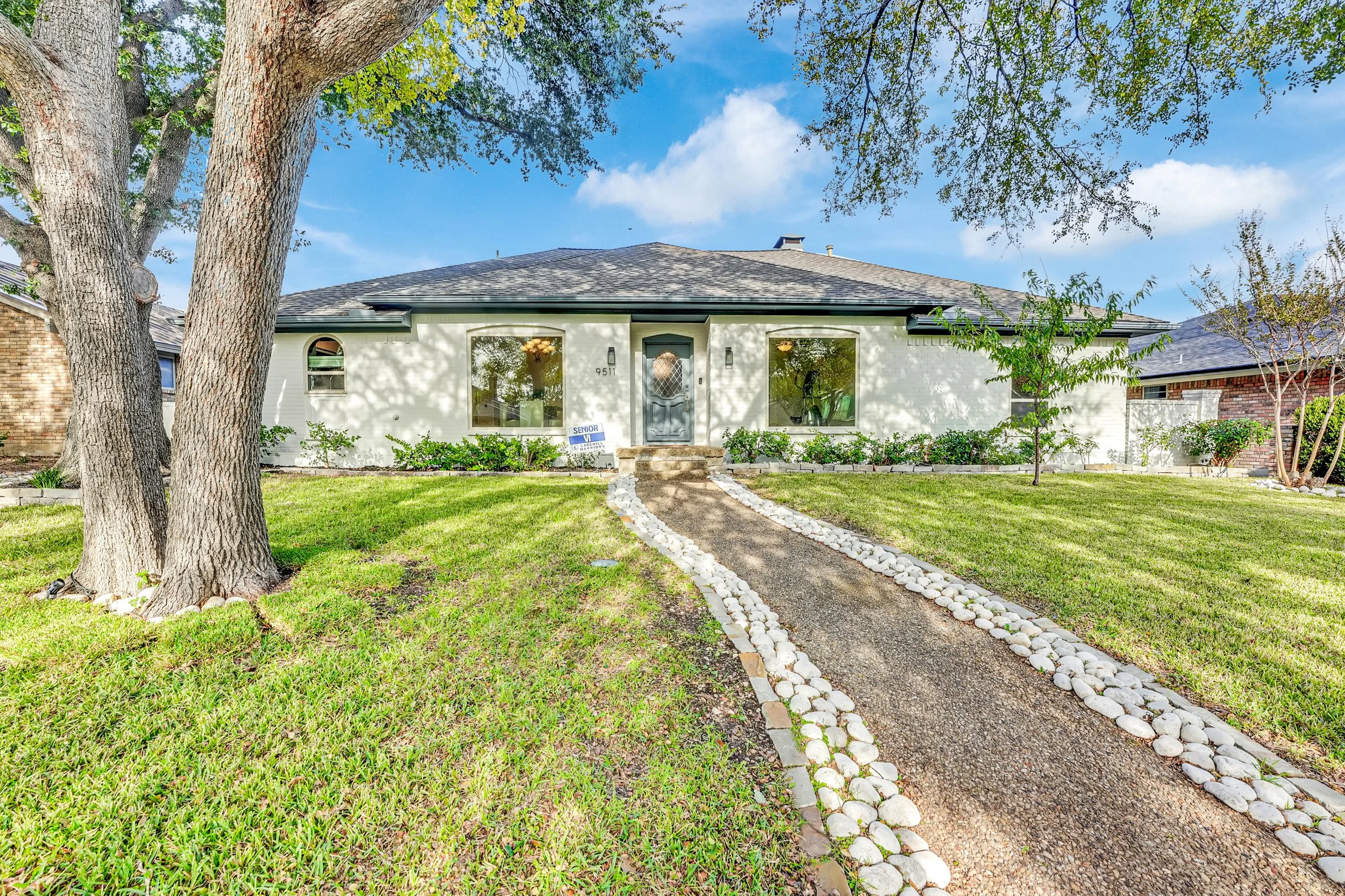 Front view of a single-story house with a gray shingled roof, white walls, and large front windows. A sidewalk with white stones on either side leads to the main door, flanked by landscaped bushes. There are trees and green grass in the front yard under a blue sky with some clouds.