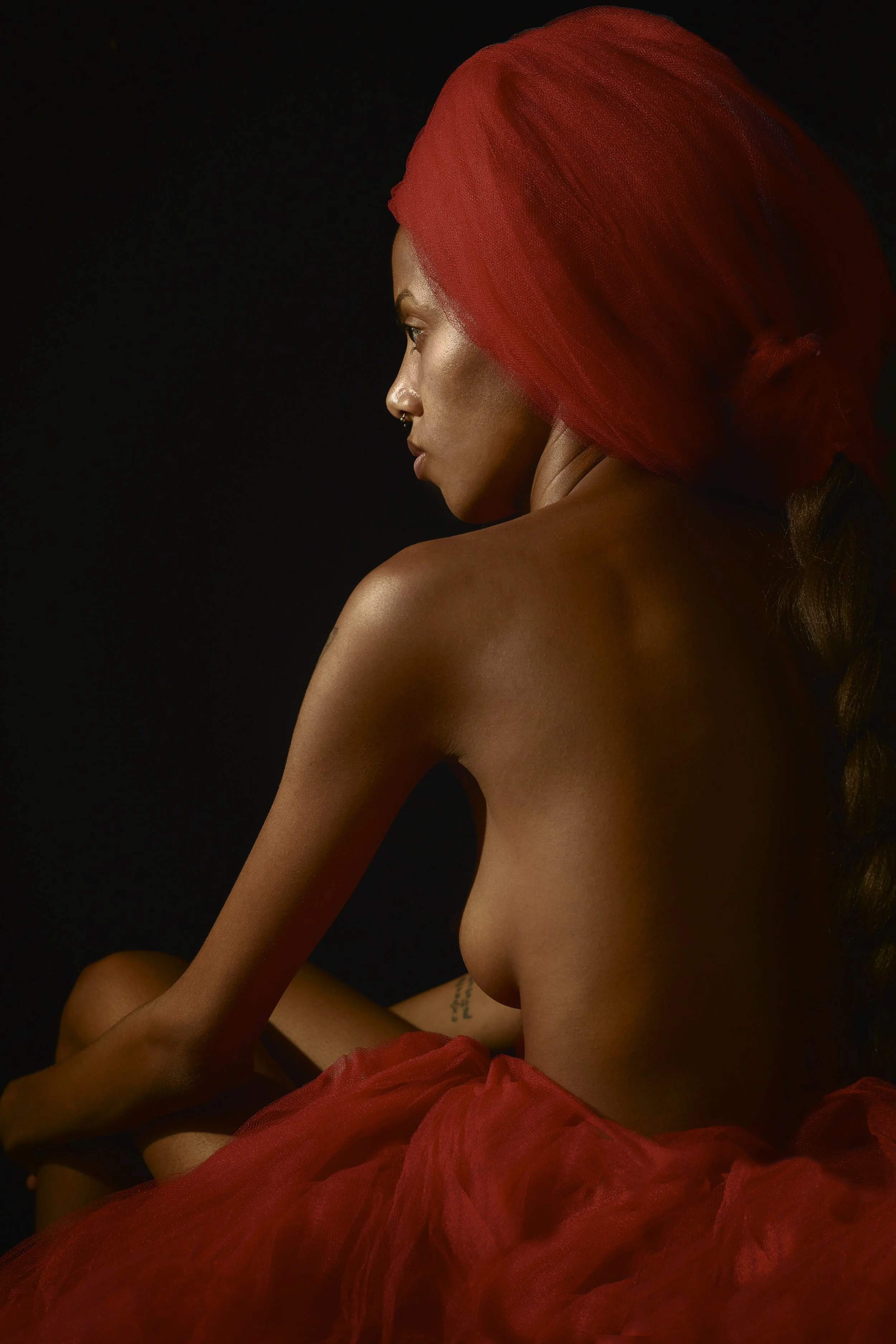 Femme nue avec un voile rouge, assise contre un fond noir, vue de profil.