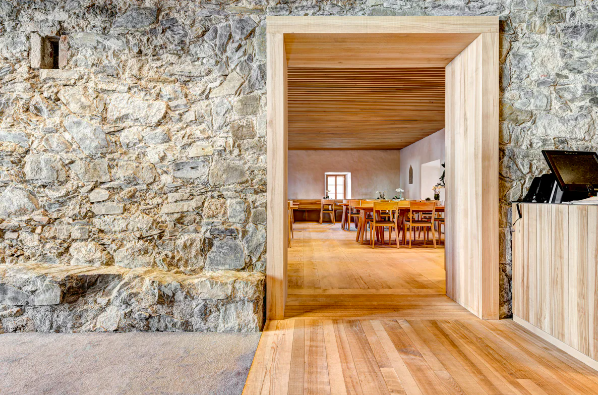 Ein Blick durch eine Tür in einen rustikalen Gemeinschaftsraum mit Holztisch und Stühlen, Holzdecke und einer kleinen Fensteröffnung.