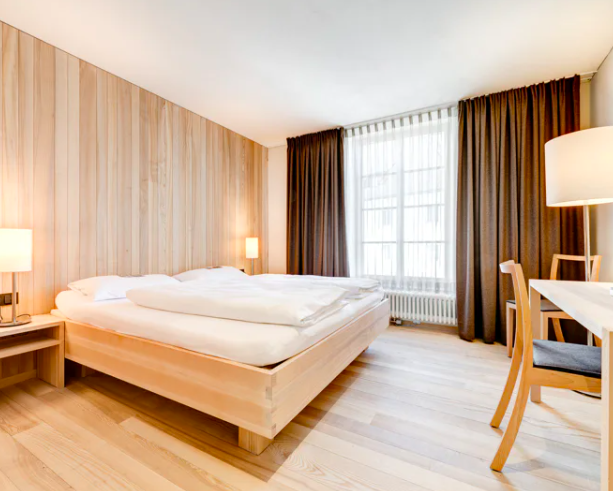 Ein Hotelzimmer mit einem großen Bett, Nachttischen mit Lampen, Holzwand, großen Fenstern mit Vorhängen, einem Schreibtisch und Stühlen.