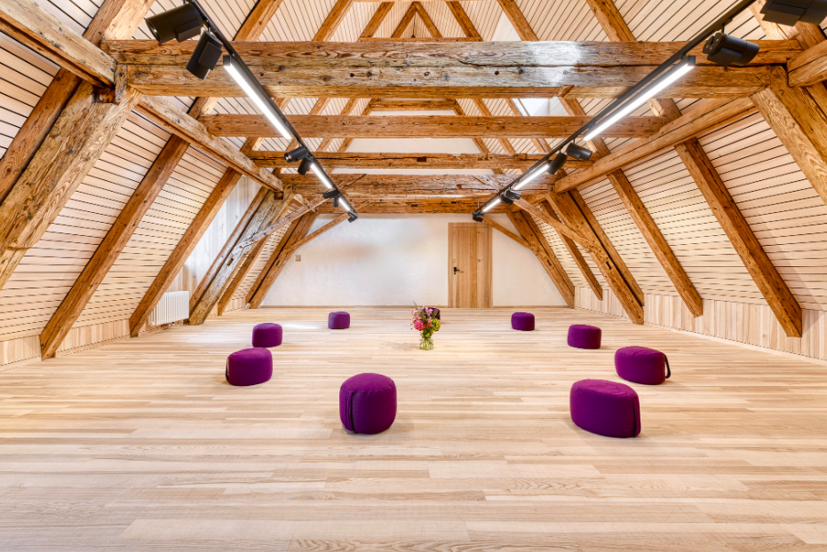 Ein heller, holzvertäfelter Dachboden mit Holzständern, einer Tür am Ende des Raumes und 8 purple Sitzkissen, die in einem Kreis um einen Blumenstrauß in einer Glasvase angeordnet sind.