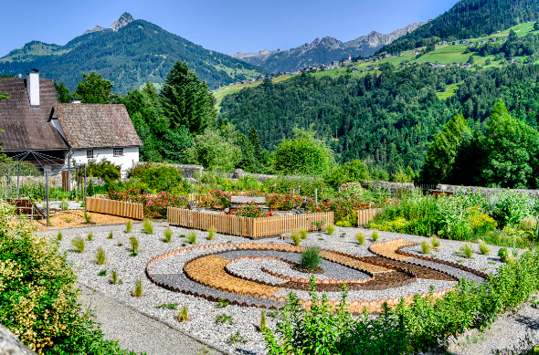 Garten mit Blumenbeeten und dekorativem Design in einer ländlichen Gegend mit Hügeln und Bergen im Hintergrund.