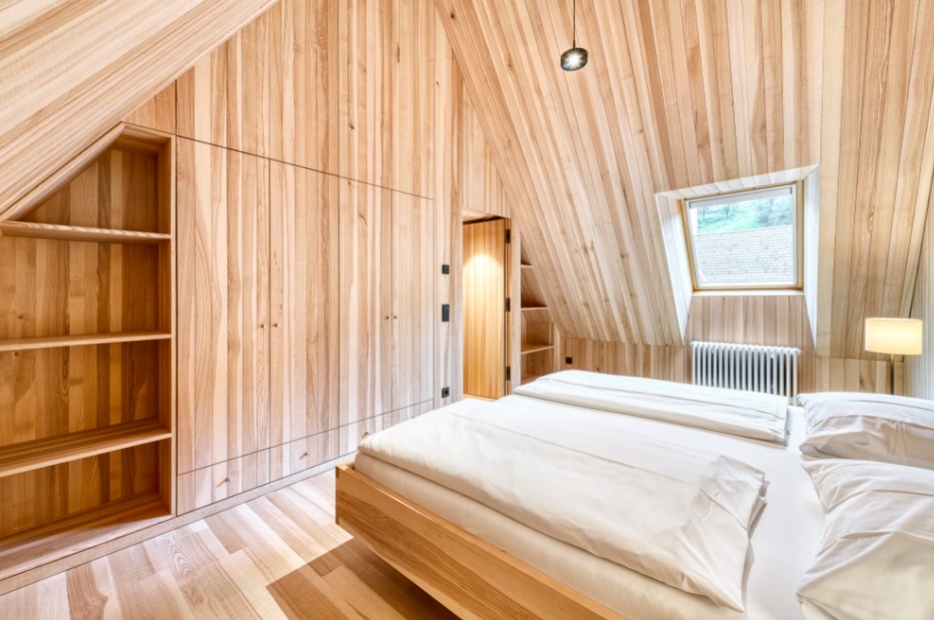 Schlafzimmer mit Holzvertäfelung, Dachschräge, Bett mit weißer Bettwäsche, Fenster an der Dachschräge, Nachttischlampe.