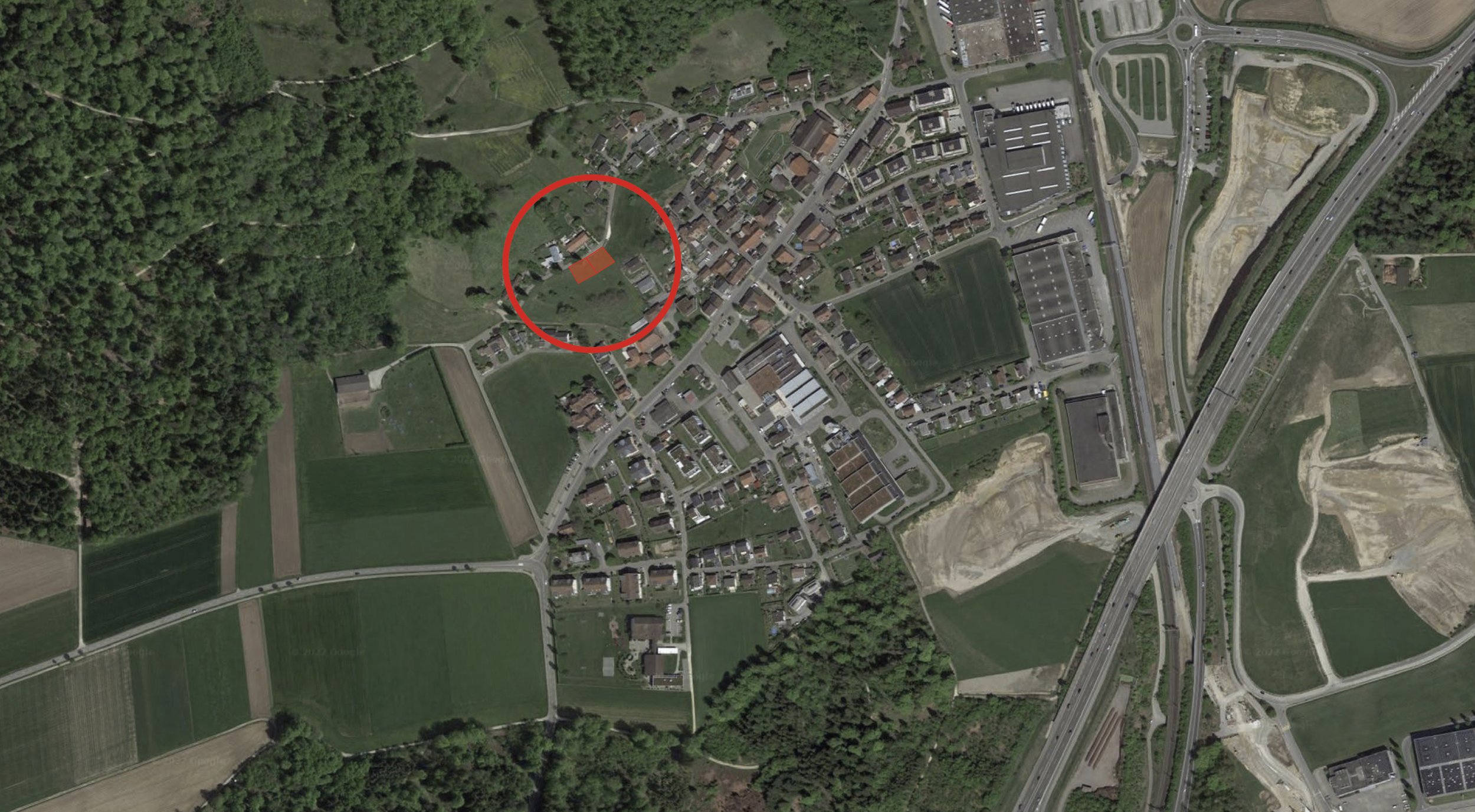 Luftaufnahme eines kleinen Dorfes mit Häusern, Feldern, Straßen und einem Waldrand im Hintergrund, mit einem roten Kreis um eine bestimmte Stelle.