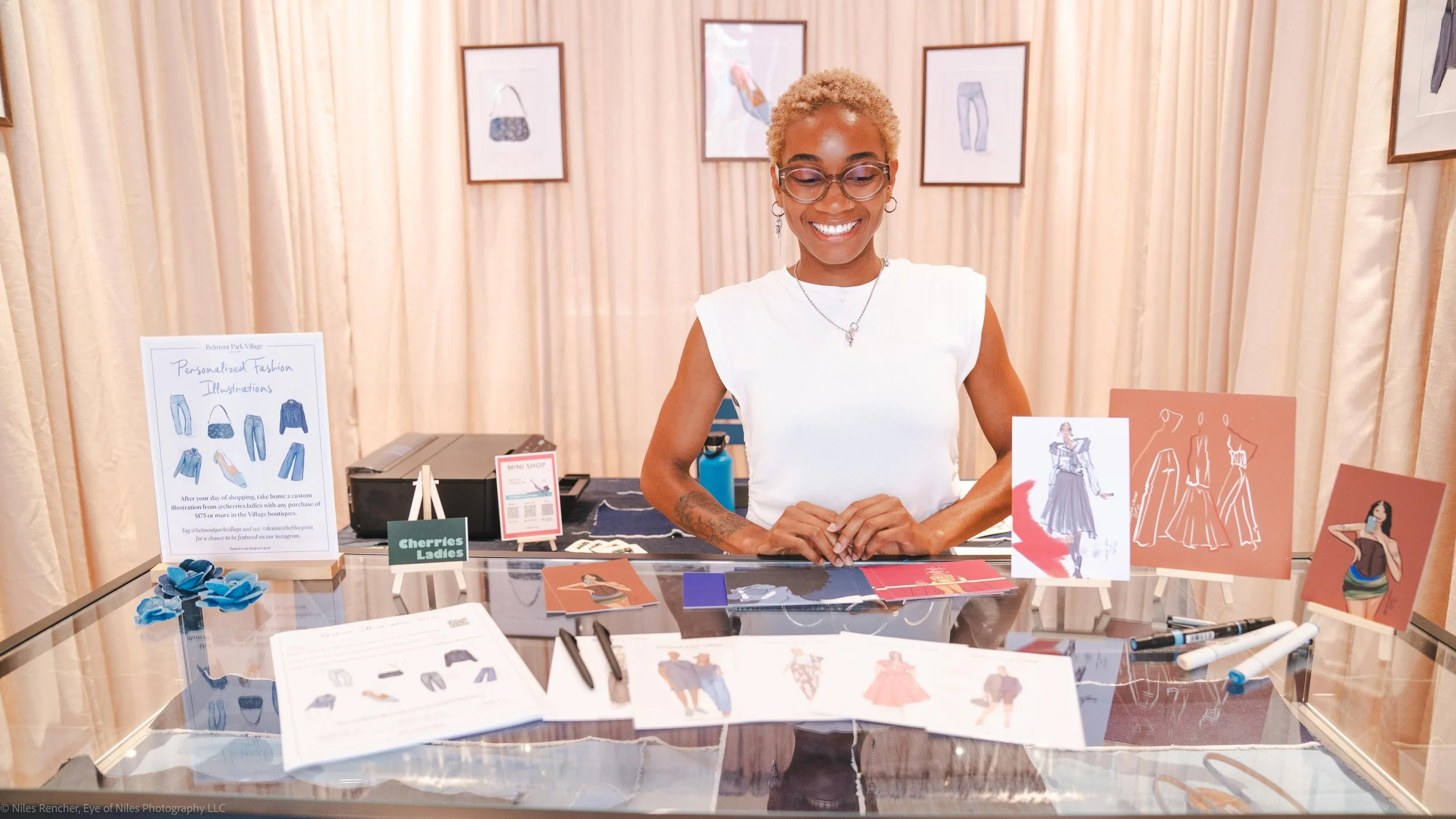 Smiling woman with short blonde curly hair and glasses sitting behind a table displaying fashion illustrations, fabric samples, and pens, with a backdrop of framed fashion sketches on cream-colored curtains. From Eye Of Niles Photography