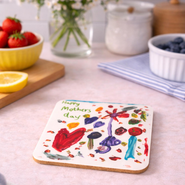 Colorful hand-painted Mother's Day card with flowers and text, placed on a pink countertop with strawberries, lemon, flowers in a glass jar, and bowls of blueberries and flowers in the background.