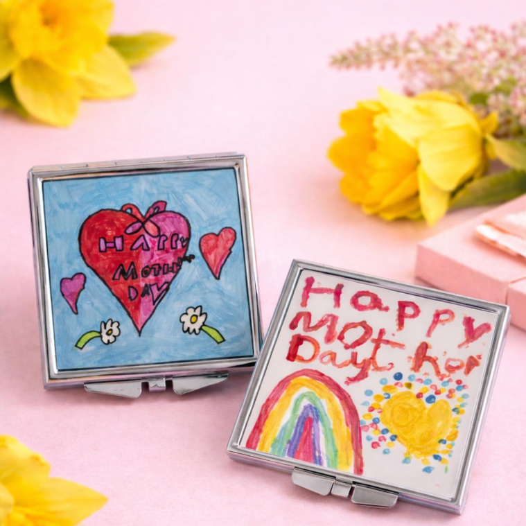 Two small mirror compacts with colorful drawings celebrating Mother's Day, surrounded by yellow flowers on a pink surface.
