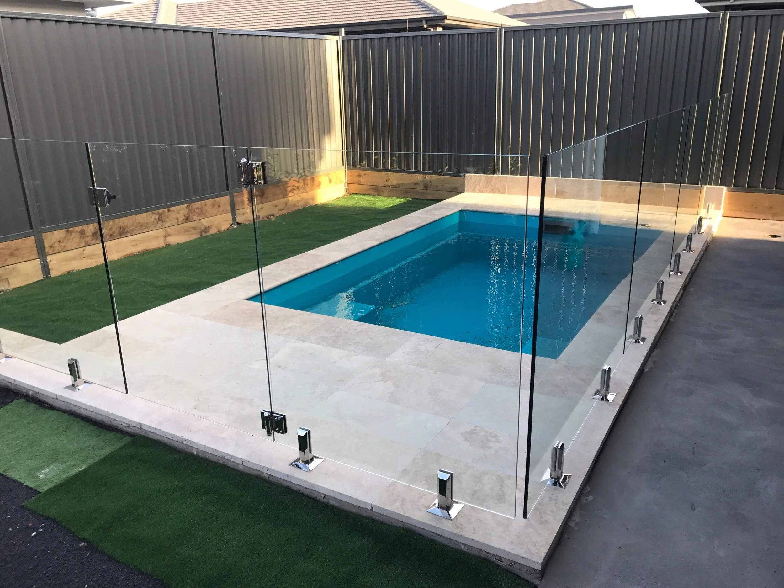 Newly installed small swimming pool with a glass safety fence in a backyard, surrounded by a grey fence and artificial grass patches.