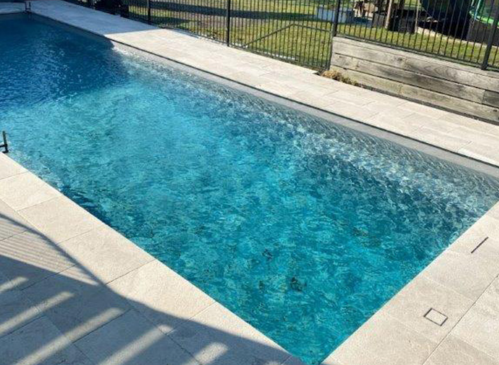 A rectangular swimming pool with light blue water, surrounded by white tiles.