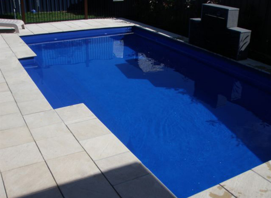A rectangular outdoor swimming pool filled with blue water, surrounded by a white tiled deck.