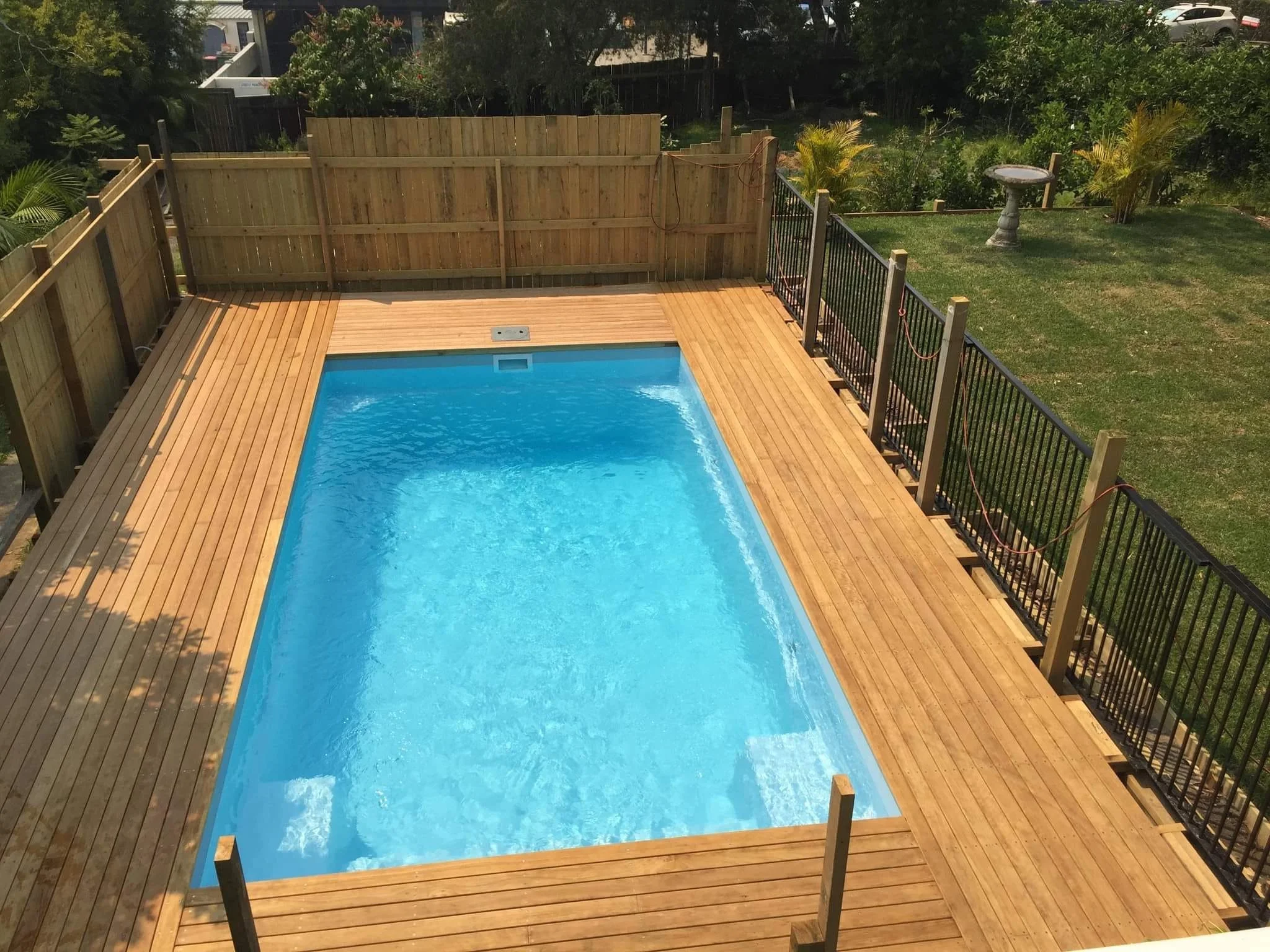 A backyard swimming pool surrounded by a wooden deck and a black metal safety fence, with a grassy lawn and some plants in the background.