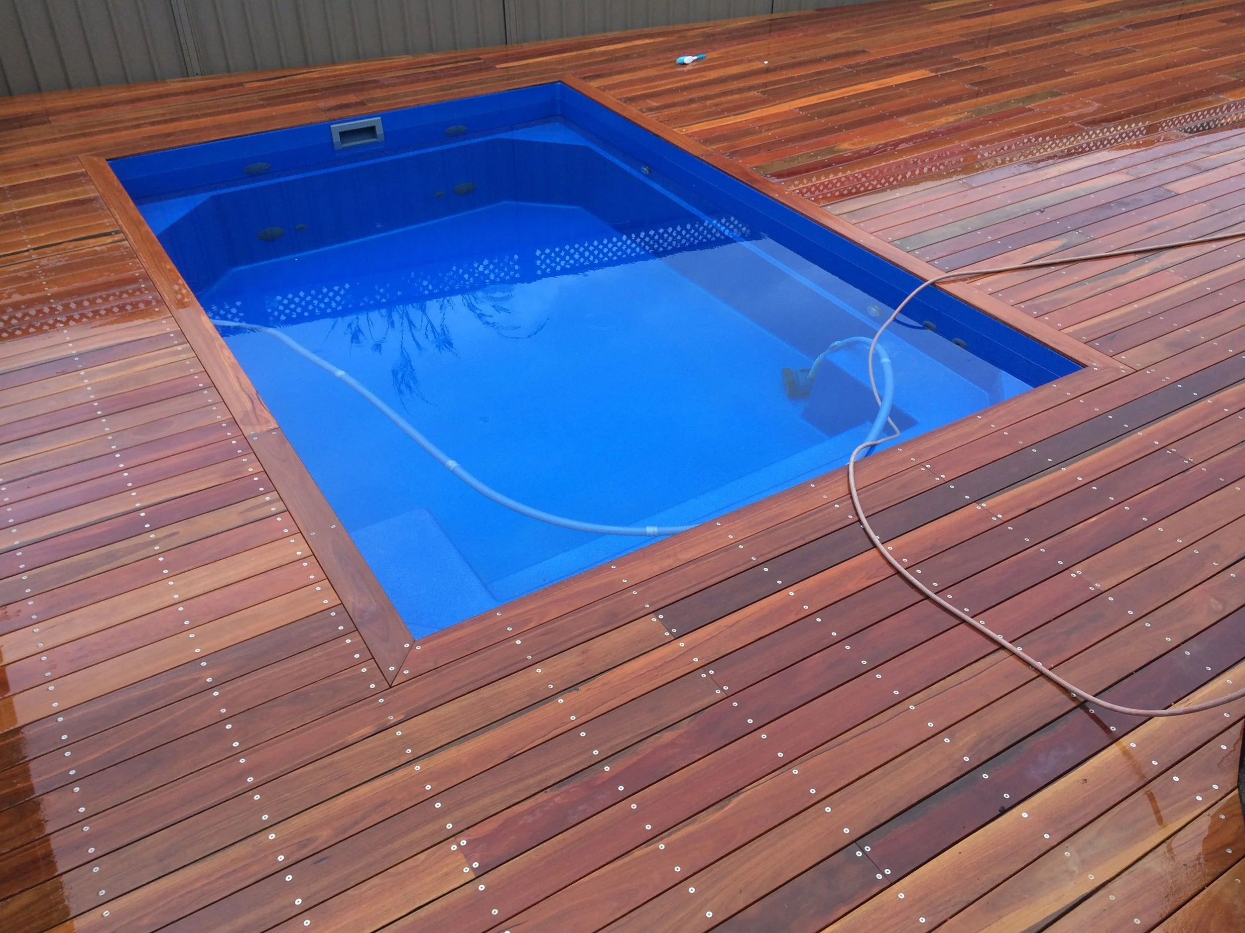 In-ground hot tub with blue interior surrounded by a wooden deck, with a hose inside.