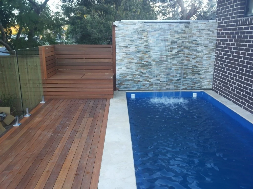 Backyard pool area with wooden deck, glass railing, a built-in wooden bench, and a water feature wall with two streams flowing into the pool, surrounded by trees and brick wall.