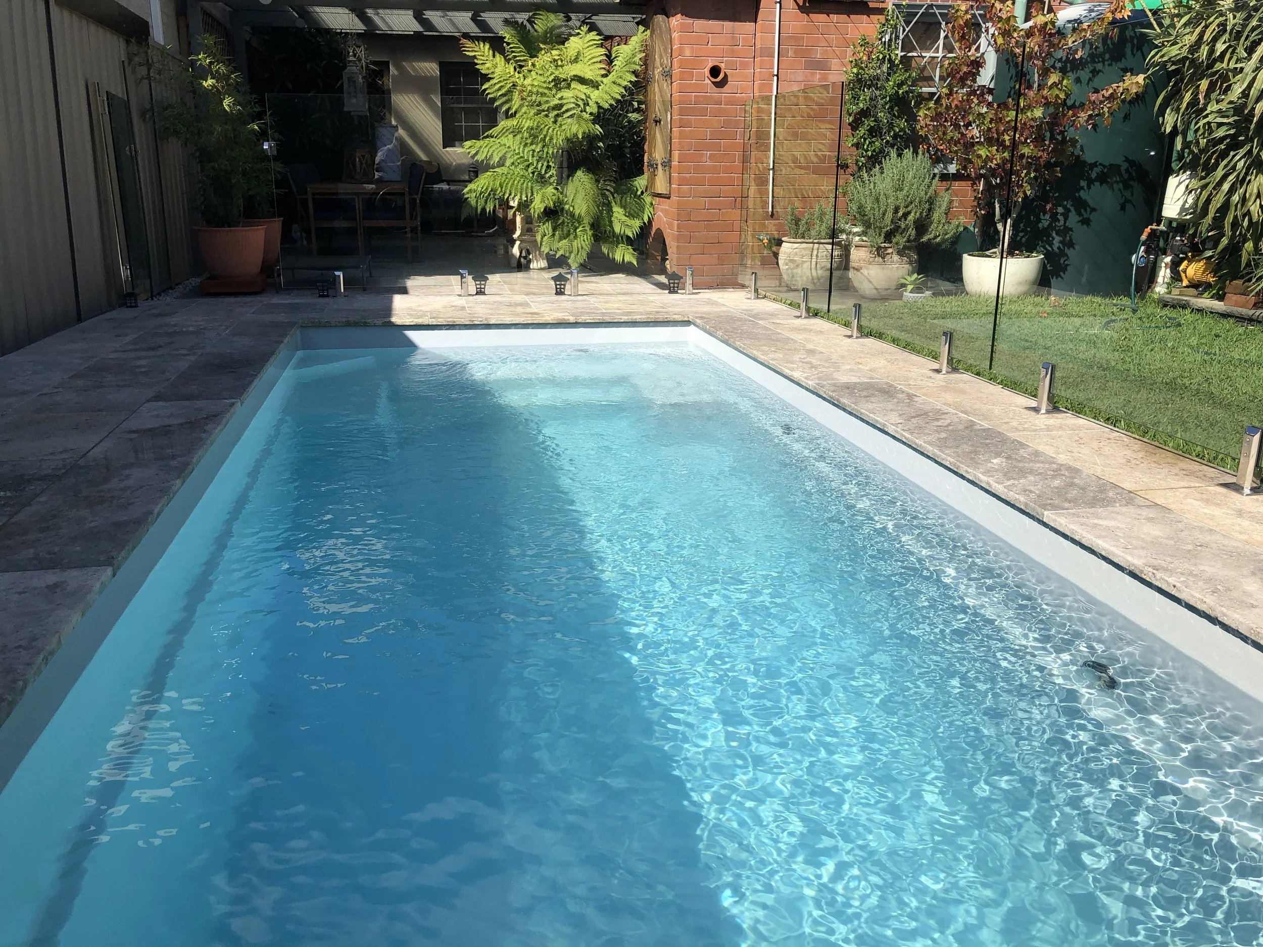 A backyard swimming pool with clear blue water, surrounded by a stone patio, potted plants, and a glass fence, with outdoor furniture and greenery in the background.