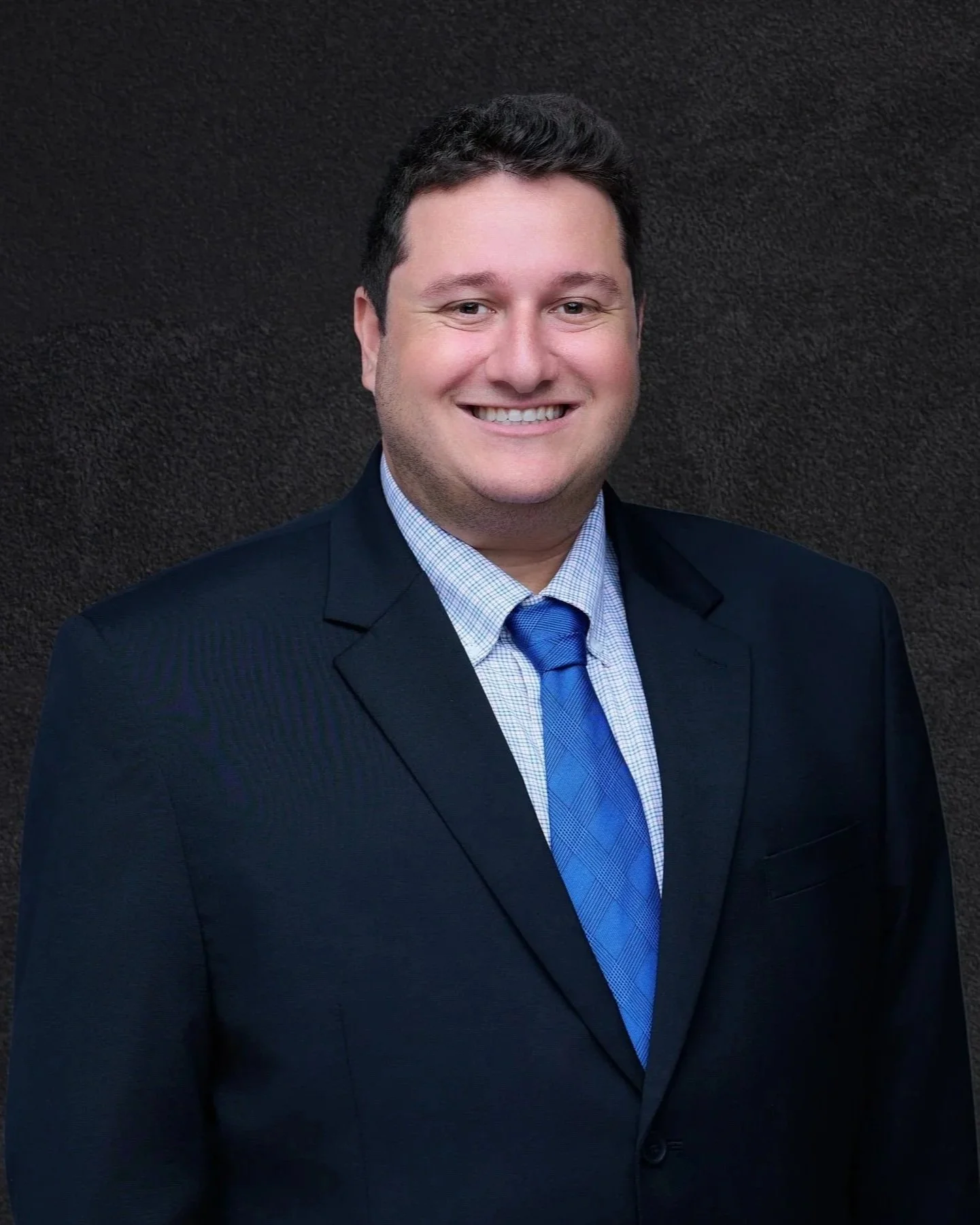 A smiling man in a dark suit, light blue shirt, and blue tie stands against a dark textured background.