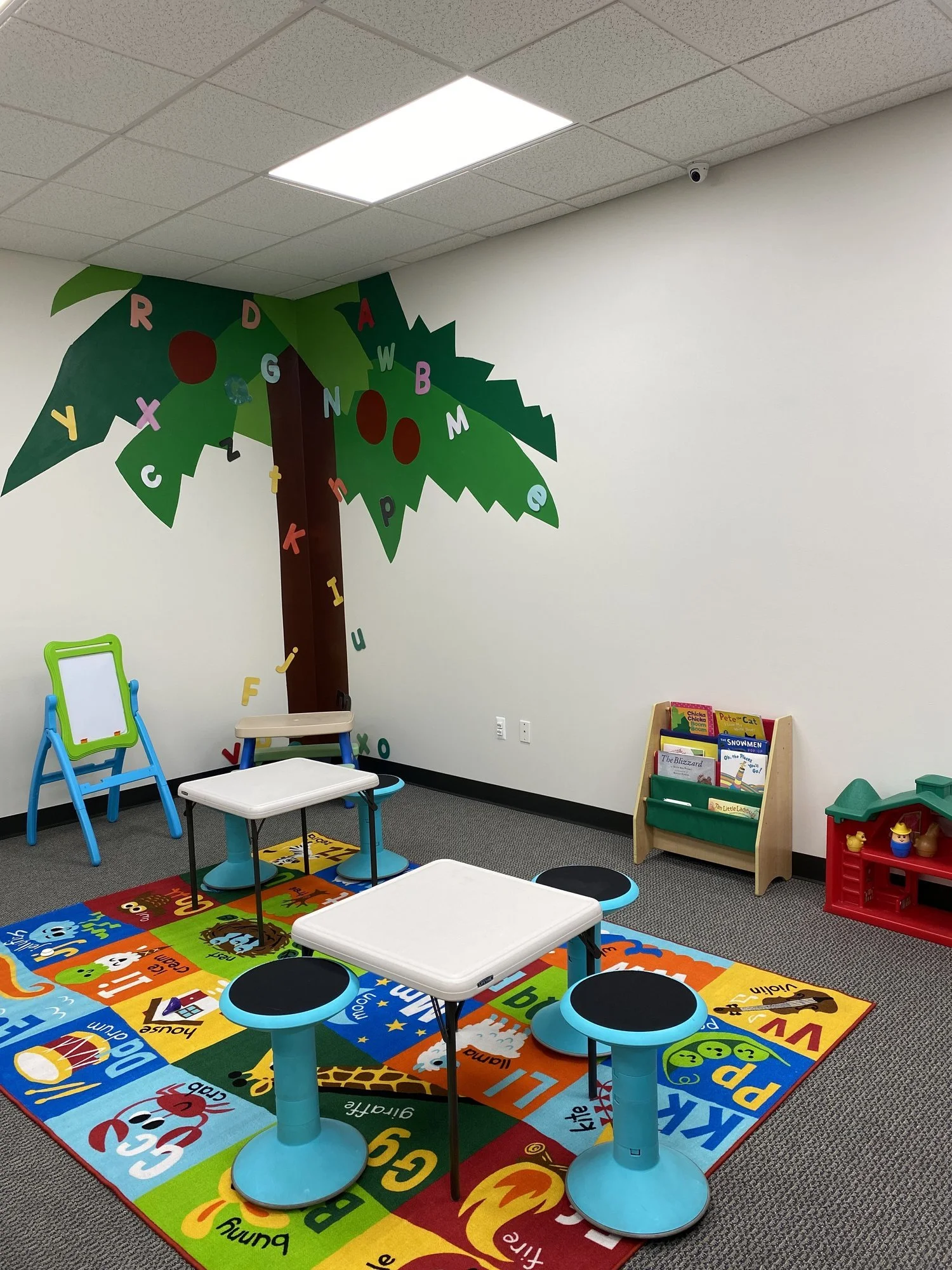 A colorful classroom with a rainbow-colored alphabet rug, three white tables with black stools, a blue and green easel, a bookshelf with children's books, a red toy shelf with yellow ducks, and a mural of a tree with alphabet letters on the wall.