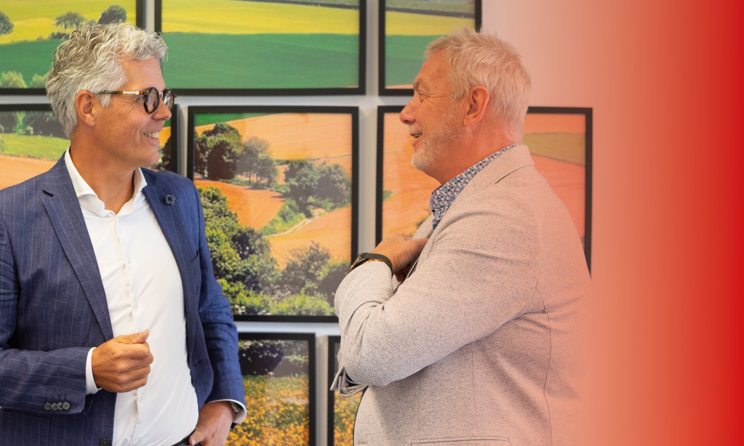 Twee mannen die lachen en praten in een kunstgalerie met landschapsfoto's op de achtergrond.