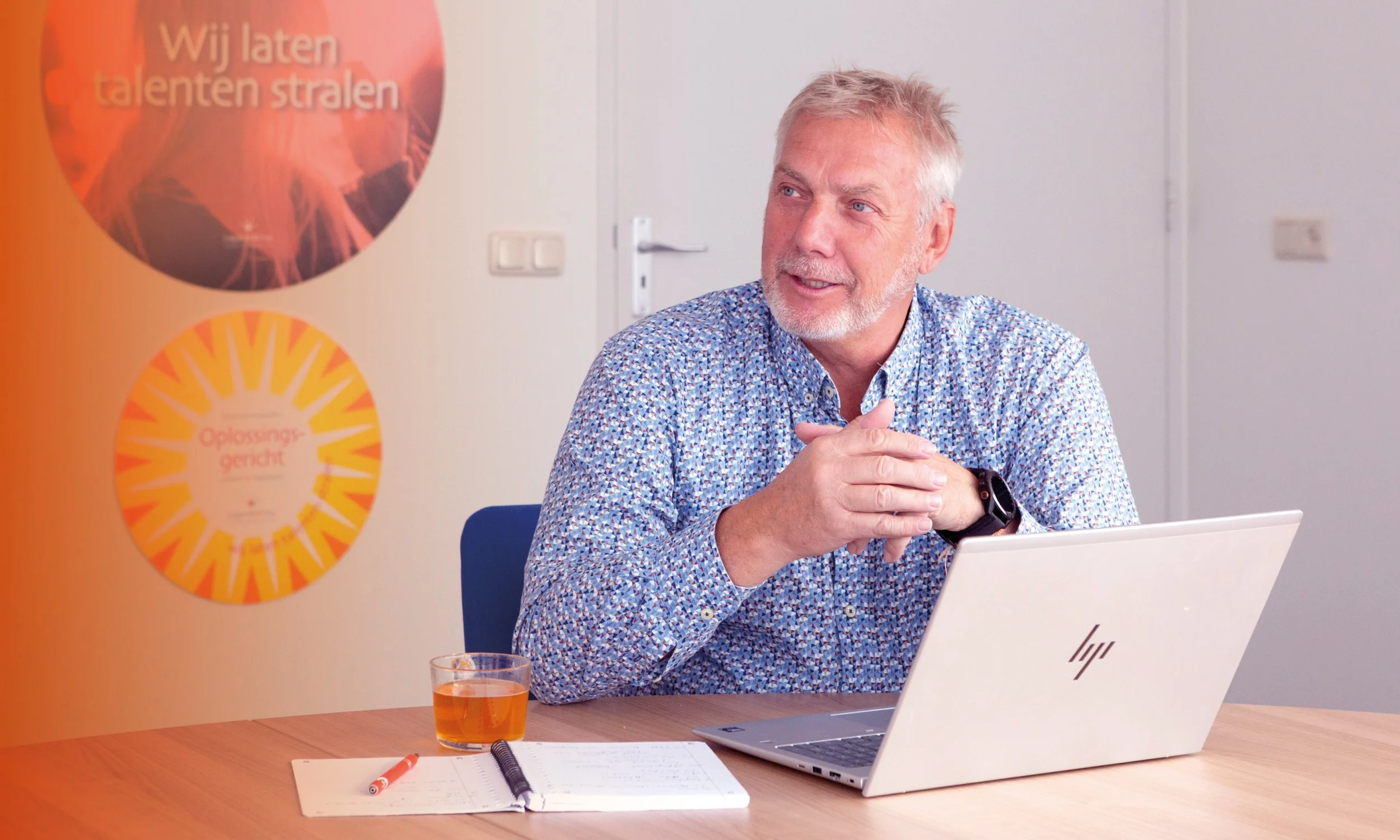 Een man met grijs haar en baard zit aan een bureau met een open HP laptop, een glas met thee, een notitieboekje en een pen. Op de muur hangen twee posters met teksten in het Nederlands.