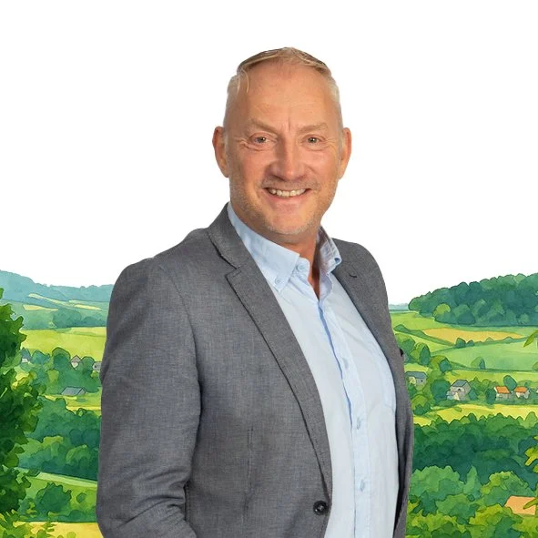 Een man met grijs haar en een lichte huid, gekleed in een grijze blazer en een lichtblauw overhemd, glimlachend voor een achtergrond van een landelijke landschap met groen landschap en heuvels.